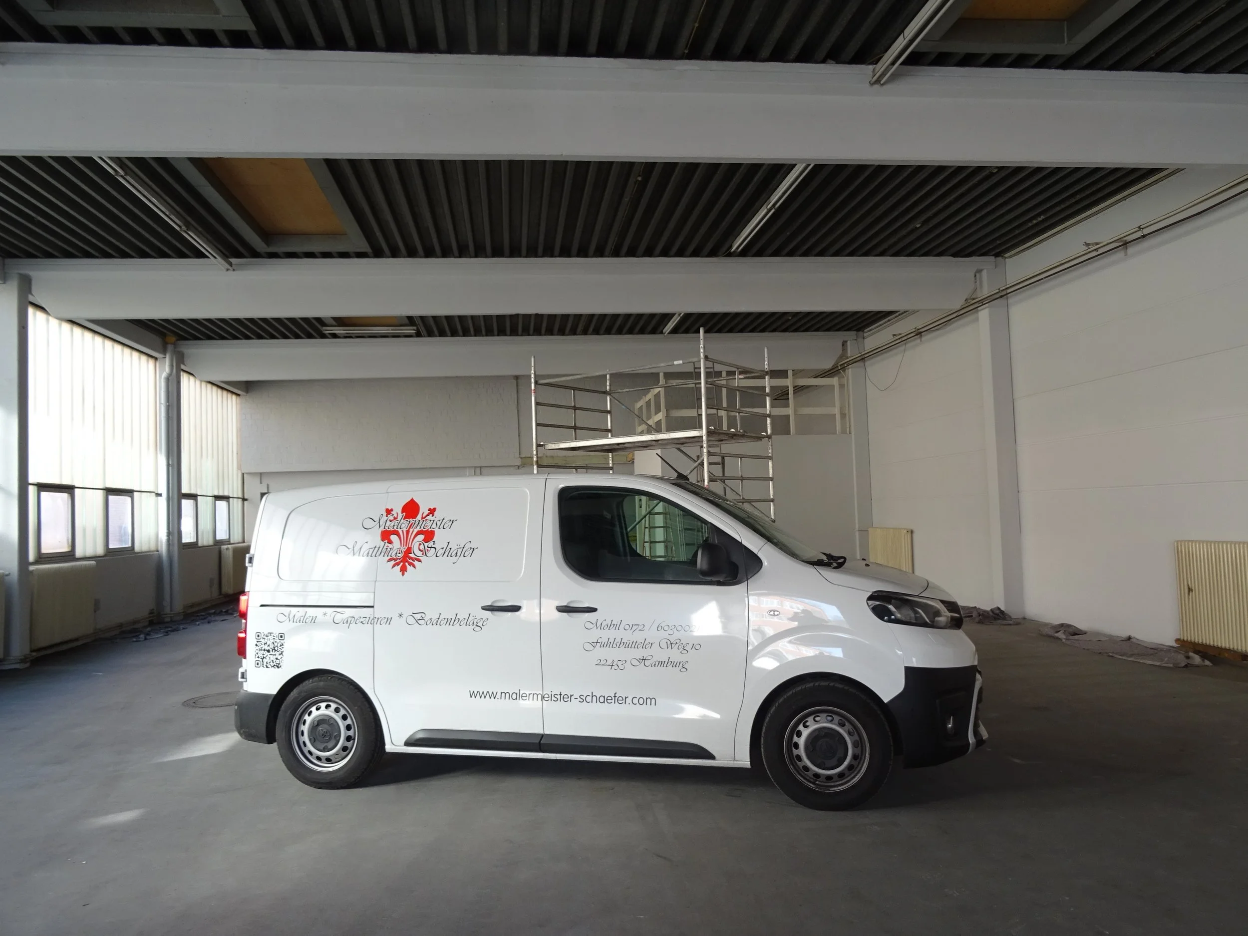 Weißer Lieferwagen mit der Aufschrift "Malermeister Matthias Schäfer" in einer leeren Halle, mit Gerüst im Hintergrund, Fenster auf der linken Seite.