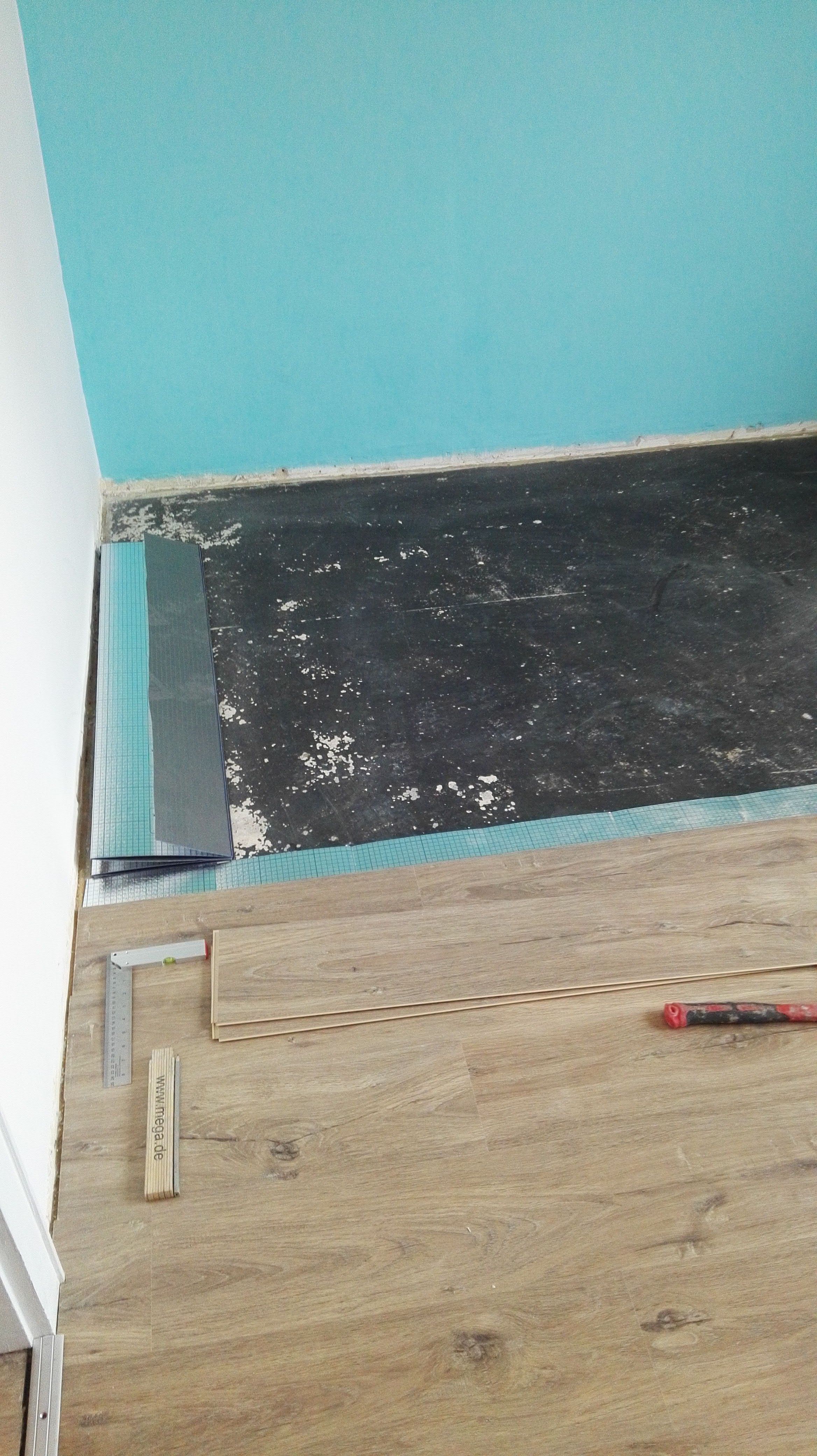 Verlegung von Laminatboden in einem Raum mit türkisfarbener Wand; Unterlage und dunkler Boden sichtbar, Werkzeuge vorhanden.