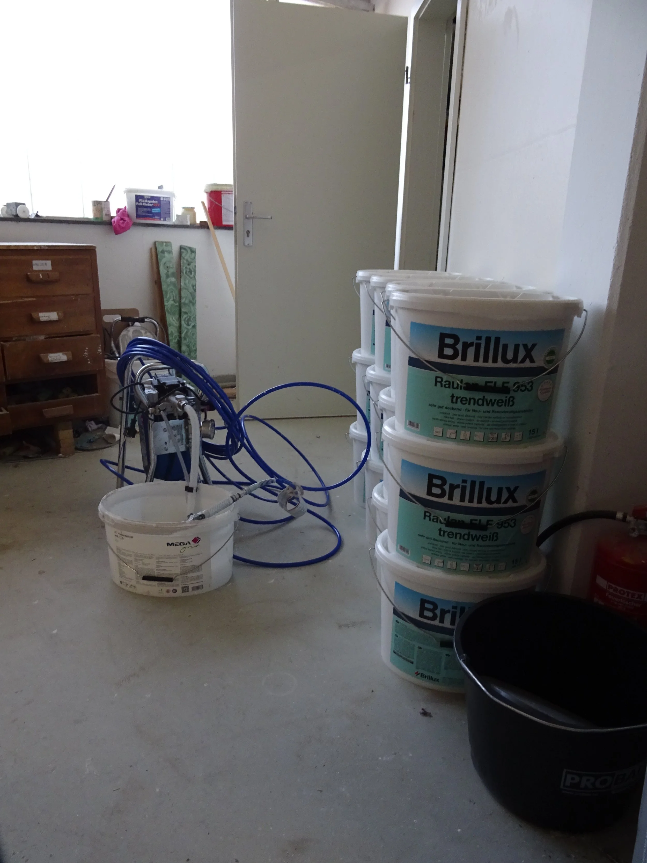 Ein Raum mit mehreren Farbeimern der Marke Brillux, einem Farbsprühgerät mit blauen Schläuchen, einer offenen Tür und einem Schrank im Hintergrund. Auf dem Boden steht ein schwarzer Eimer und ein weiterer Farbeimer. Der Raum scheint als Lager für Mal