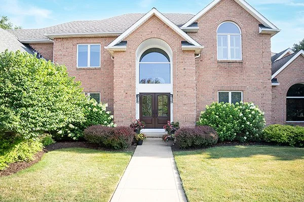 Front view of a two-story brick house with a curved arched window above the dark wooden front door, surrounded by green shrubs and flowering plants, with a concrete pathway leading to the entrance.