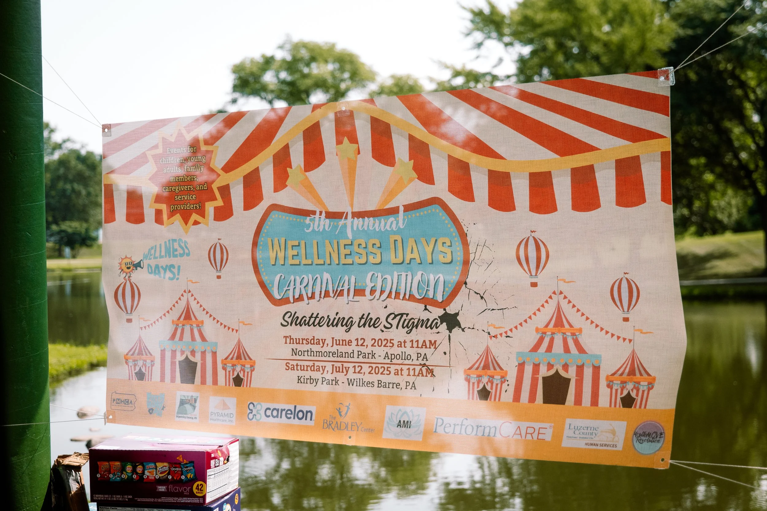 A colorful carnival-themed banner advertising the 5th Annual Wellness Days Carnival Edition, titled "Shattering the Stigma," scheduled for June 12 and July 12, 2025, at 11 AM, held at Northmoreland Park in Apollo, PA, and Kirby Park in Wilkes Barre, PA. The banner features circus tents, balloons, and a striped canopy.