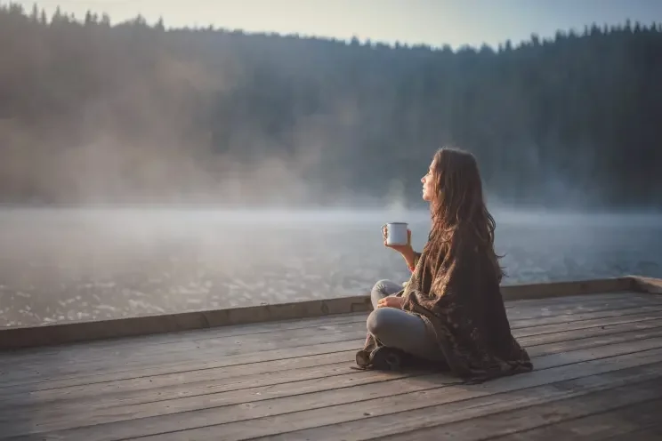 A woman sitting cross-legged on a wooden dock by a lake, holding a mug, with mist rising over the water and a forest in the background.