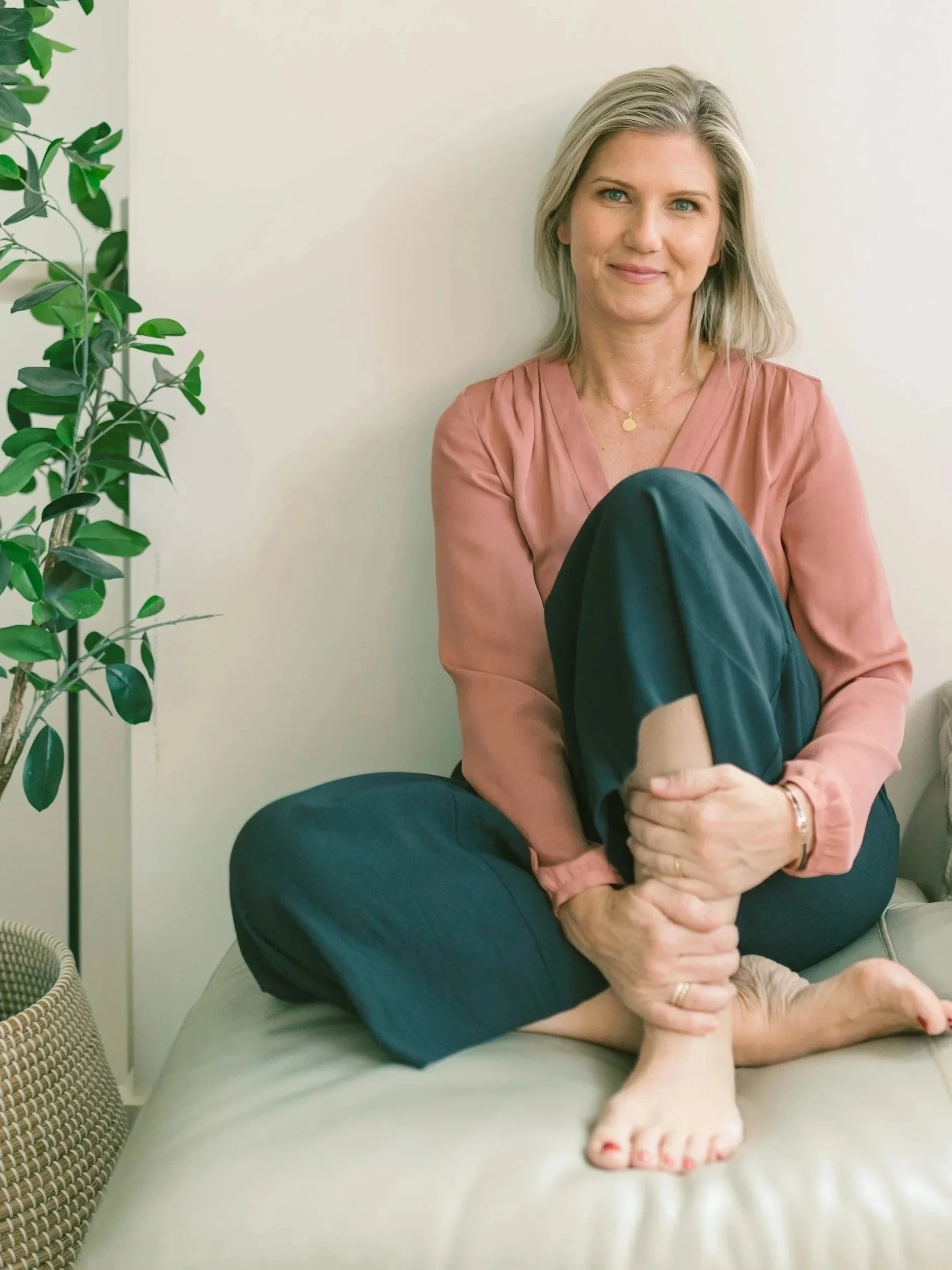 A woman sitting on a sofa, holding her shin. She is smiling and has light gray hair, wearing a pink blouse and dark pants, with a plant nearby.