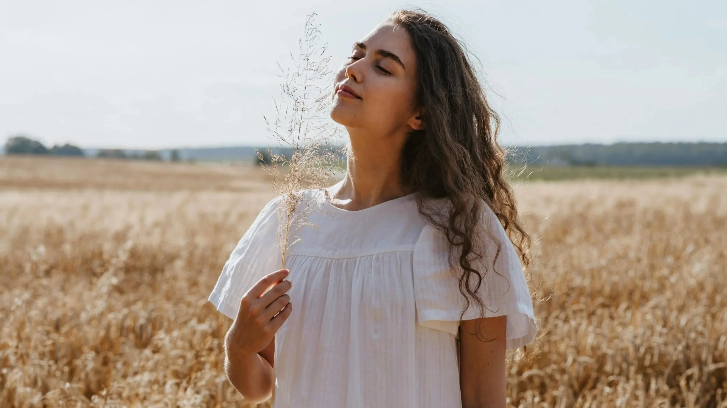 A young woman with long, curly brown hair stands in a golden wheat field, holding a small sprig of wheat, with eyes closed and a peaceful expression.