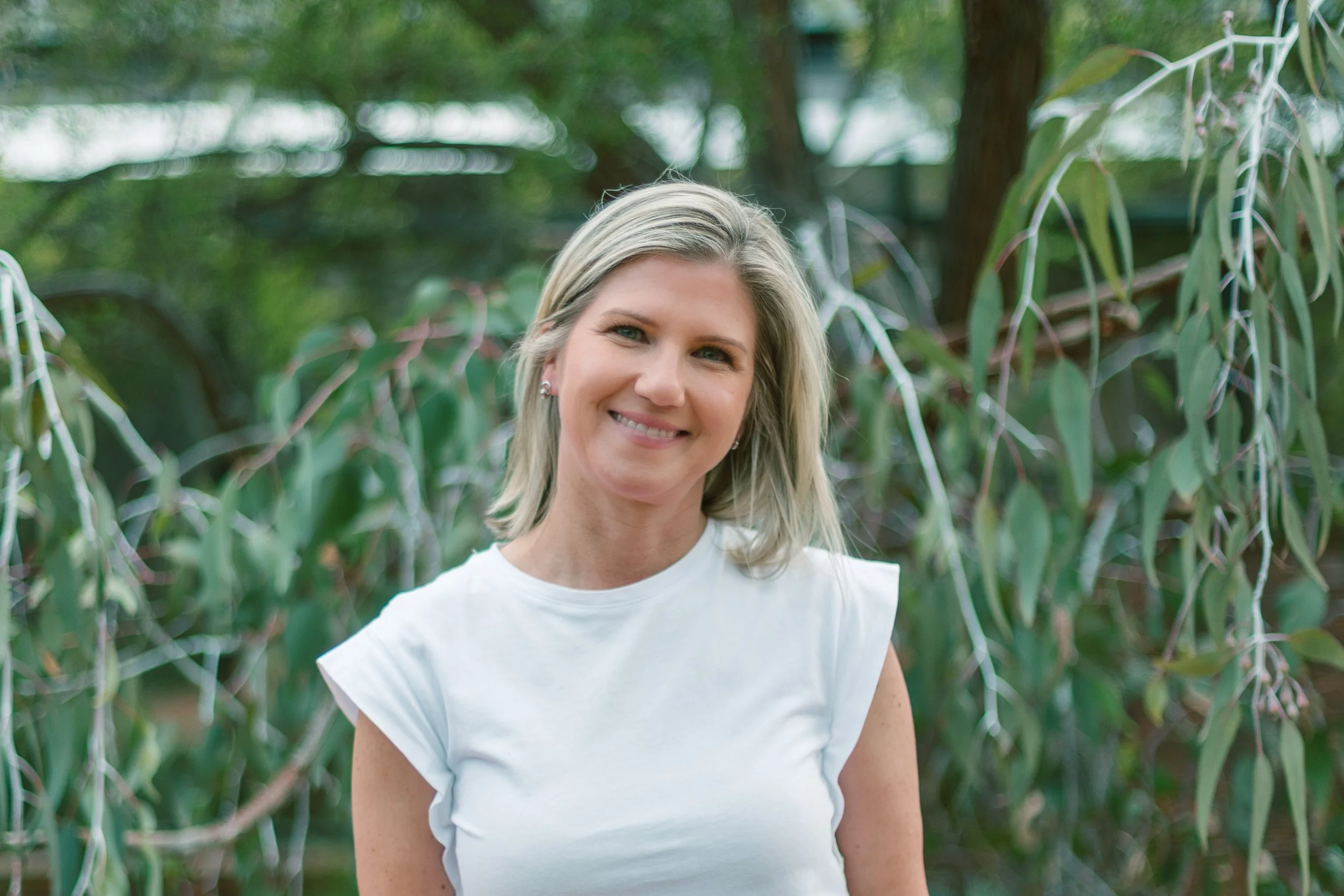 A smiling woman with blonde hair wearing a white sleeveless top standing outdoors with leafy green trees in the background.