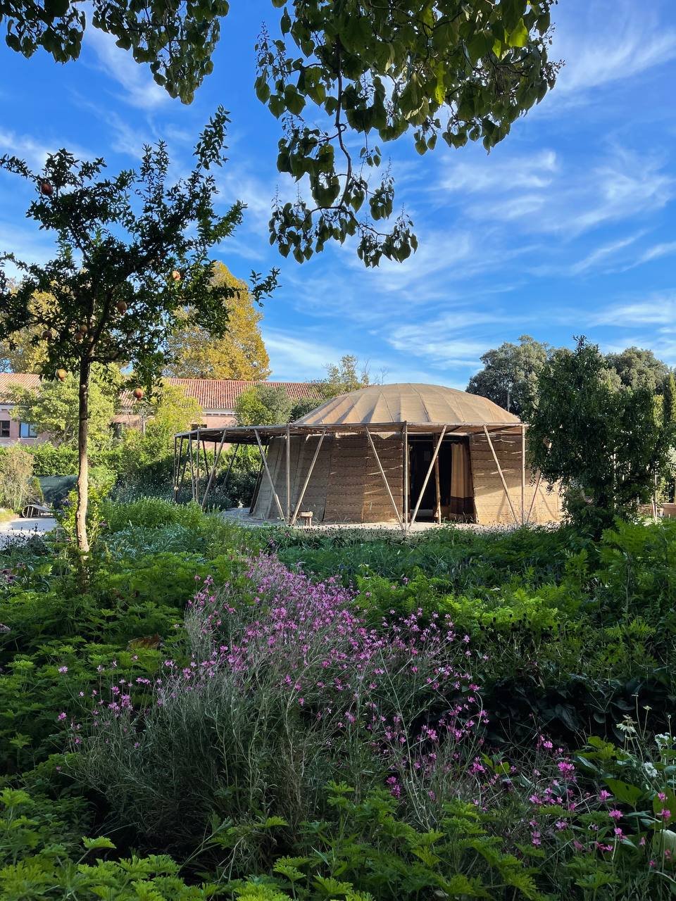 Circular wooden structure with a domed roof surrounded by lush green garden and pink flowers, set against a clear blue sky and trees.