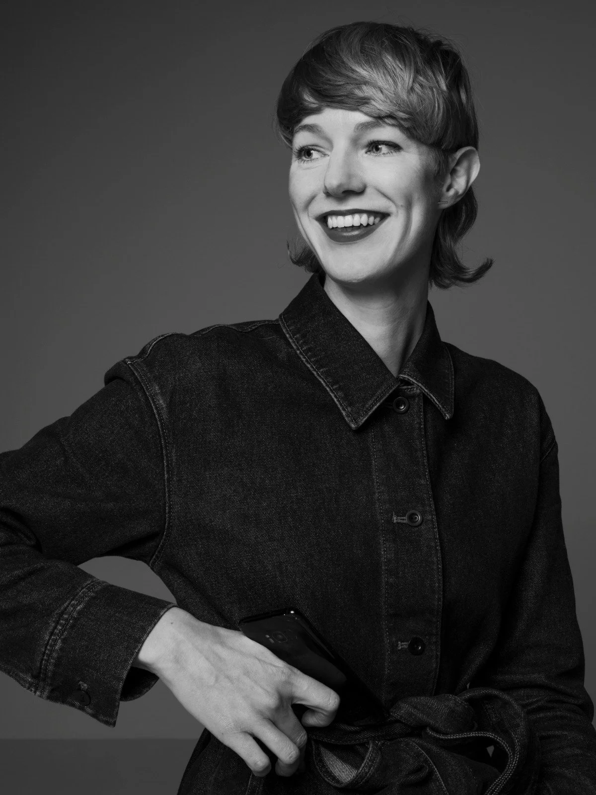 Black and white portrait of a smiling person with short hair wearing a denim jacket, holding a smartphone.