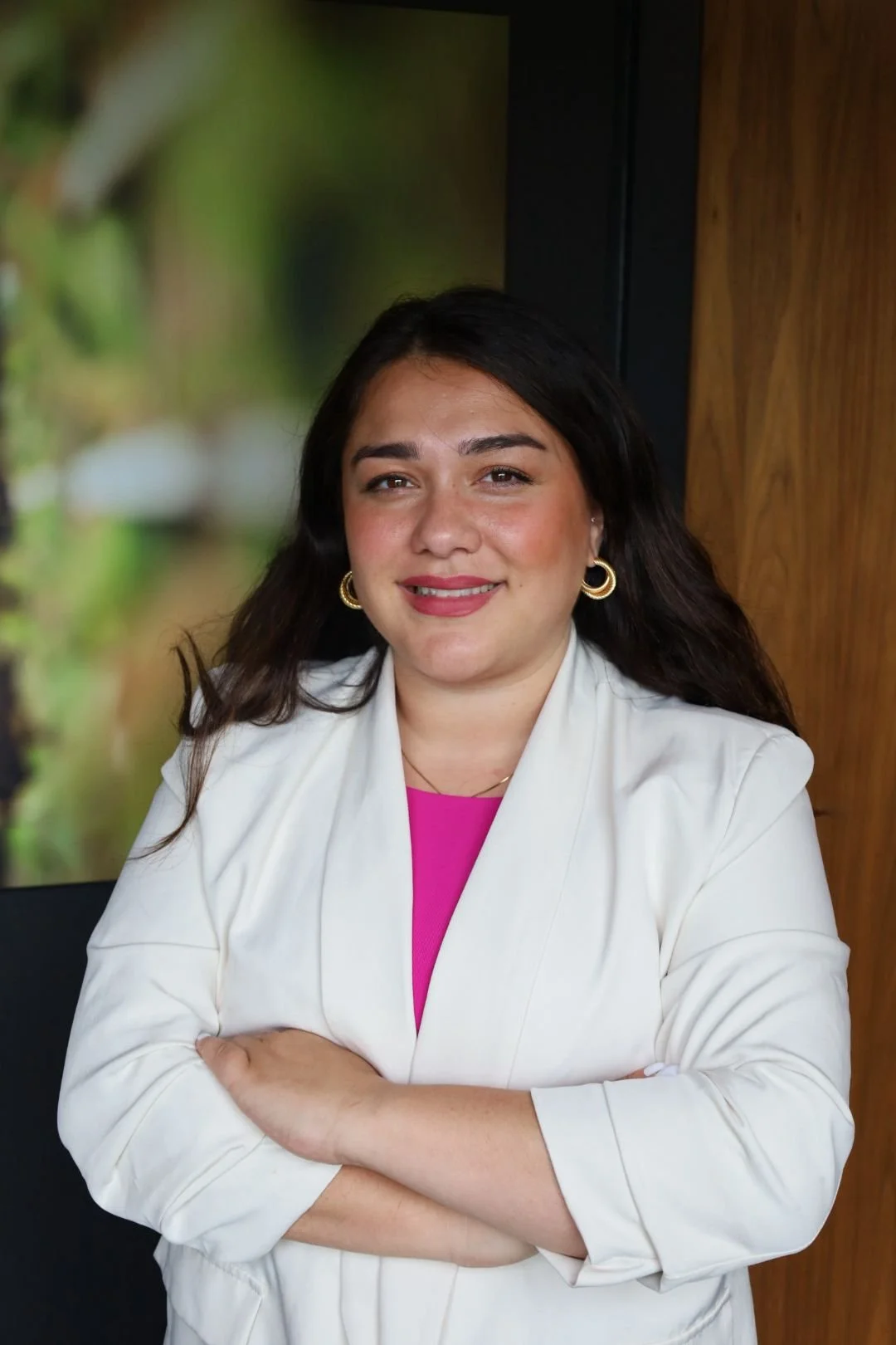 A woman with shoulder-length black hair smiling at the camera, wearing a black blazer and a black shirt, with a blurred colorful artwork in the background.