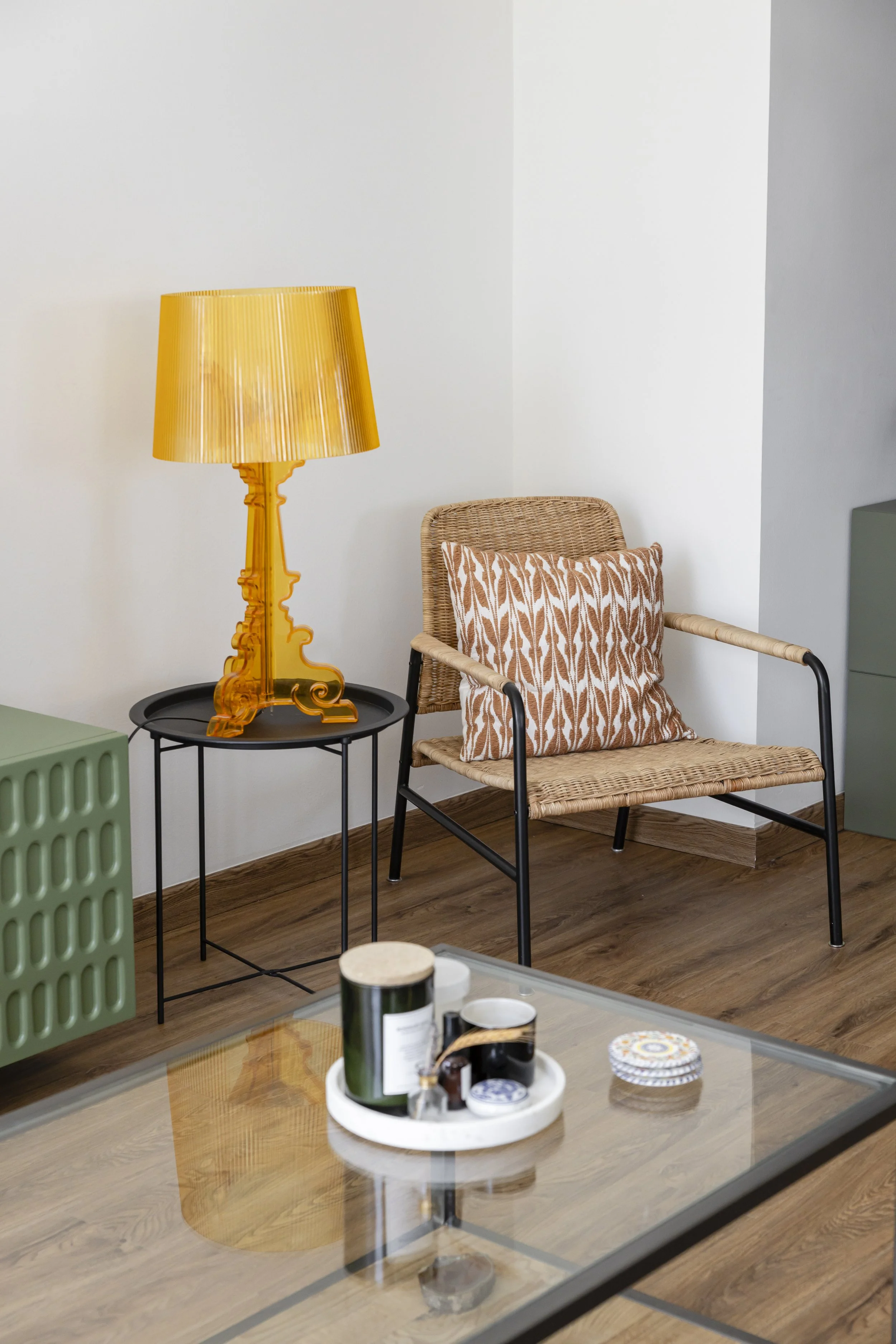 A living room corner with a wicker chair that has a decorative pillow, a side table with a yellow lamp, and a glass coffee table with a tray of candles and small items.