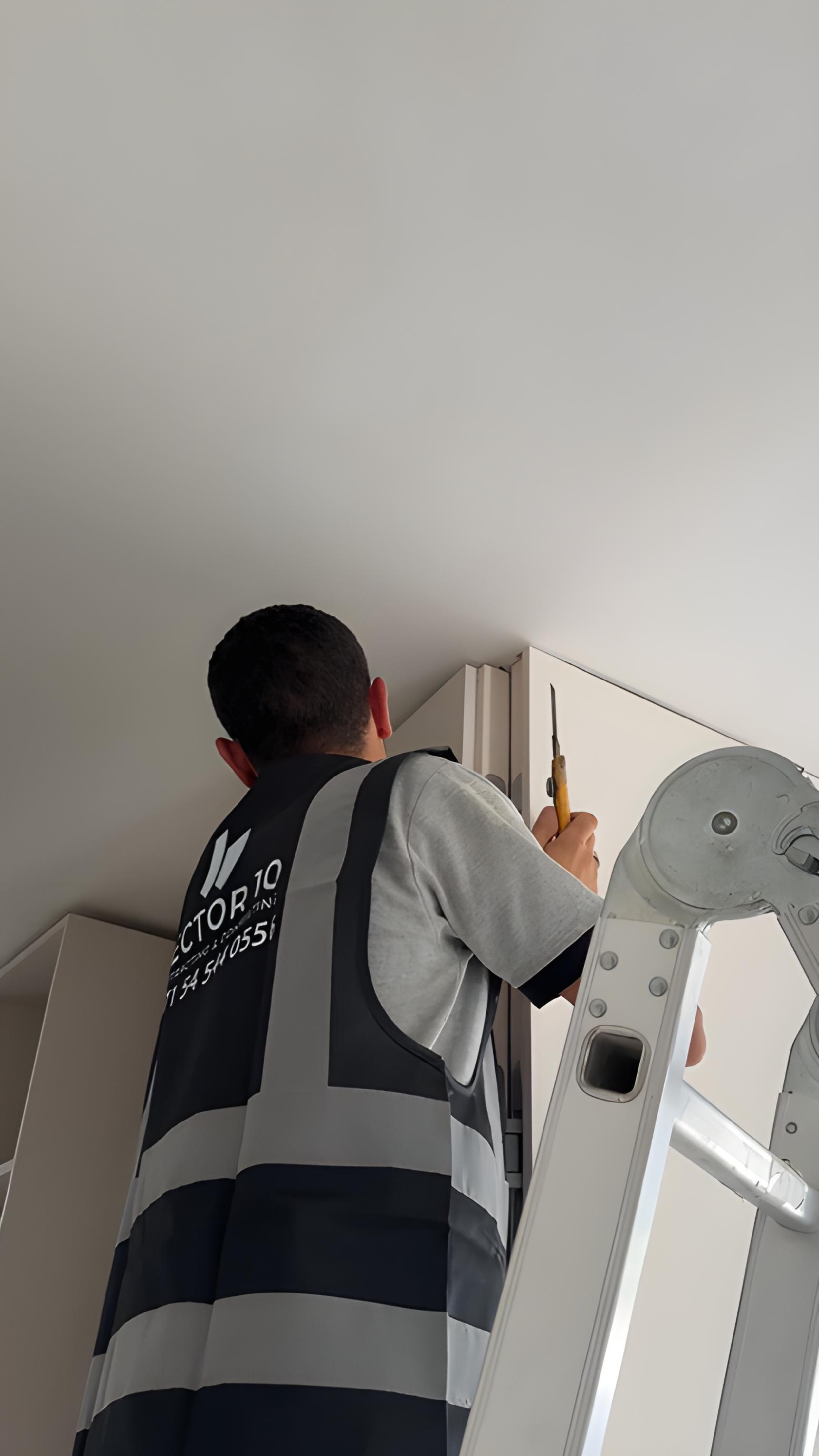 A person in a grey shirt and black safety vest standing on a ladder, working on overhead cabinets with a screwdriver.