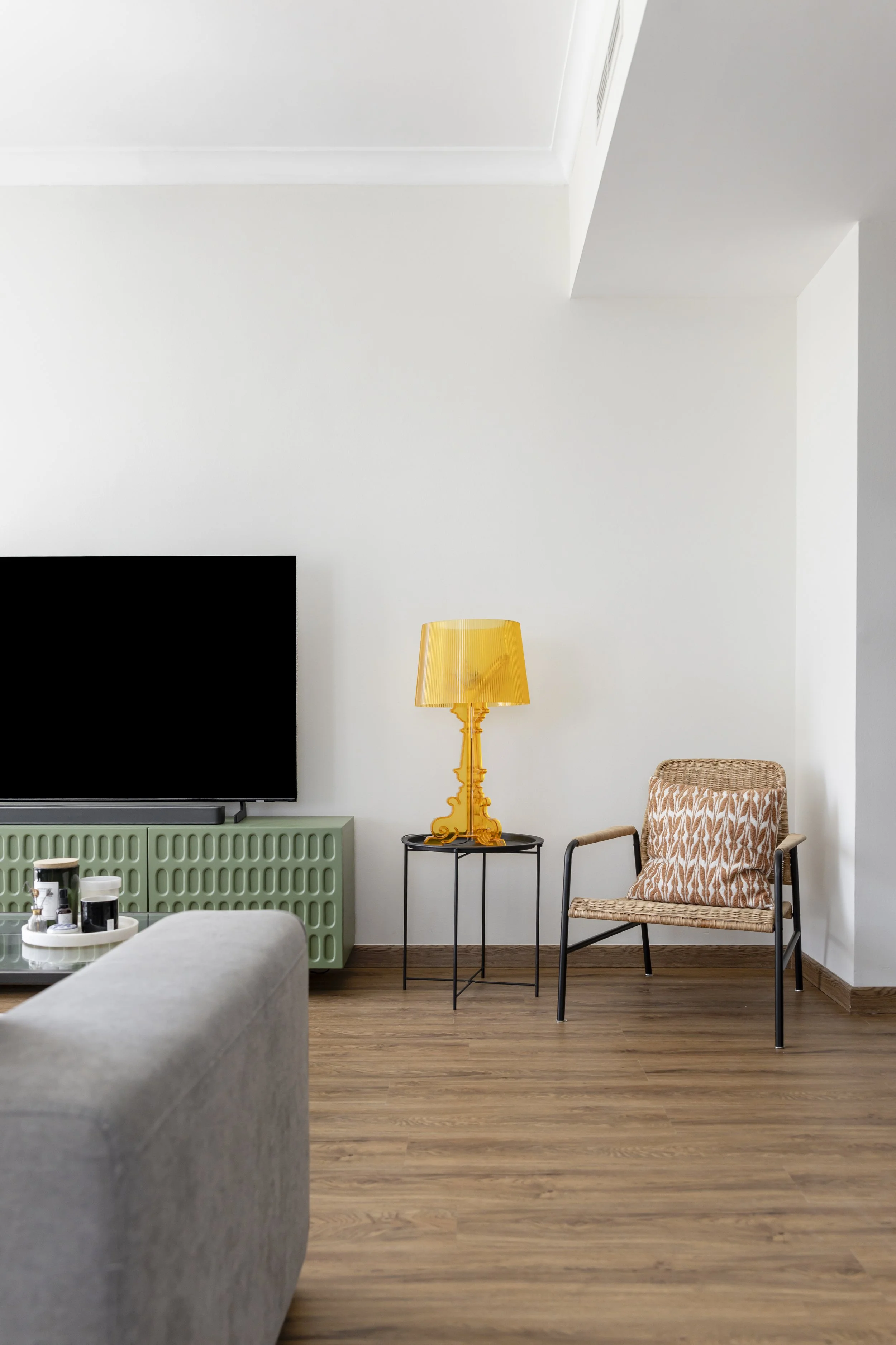 Living room with a gray sofa, a black TV on a green radiator cover, a yellow lamp on a small black table, a woven chair with a patterned pillow, and hardwood flooring.