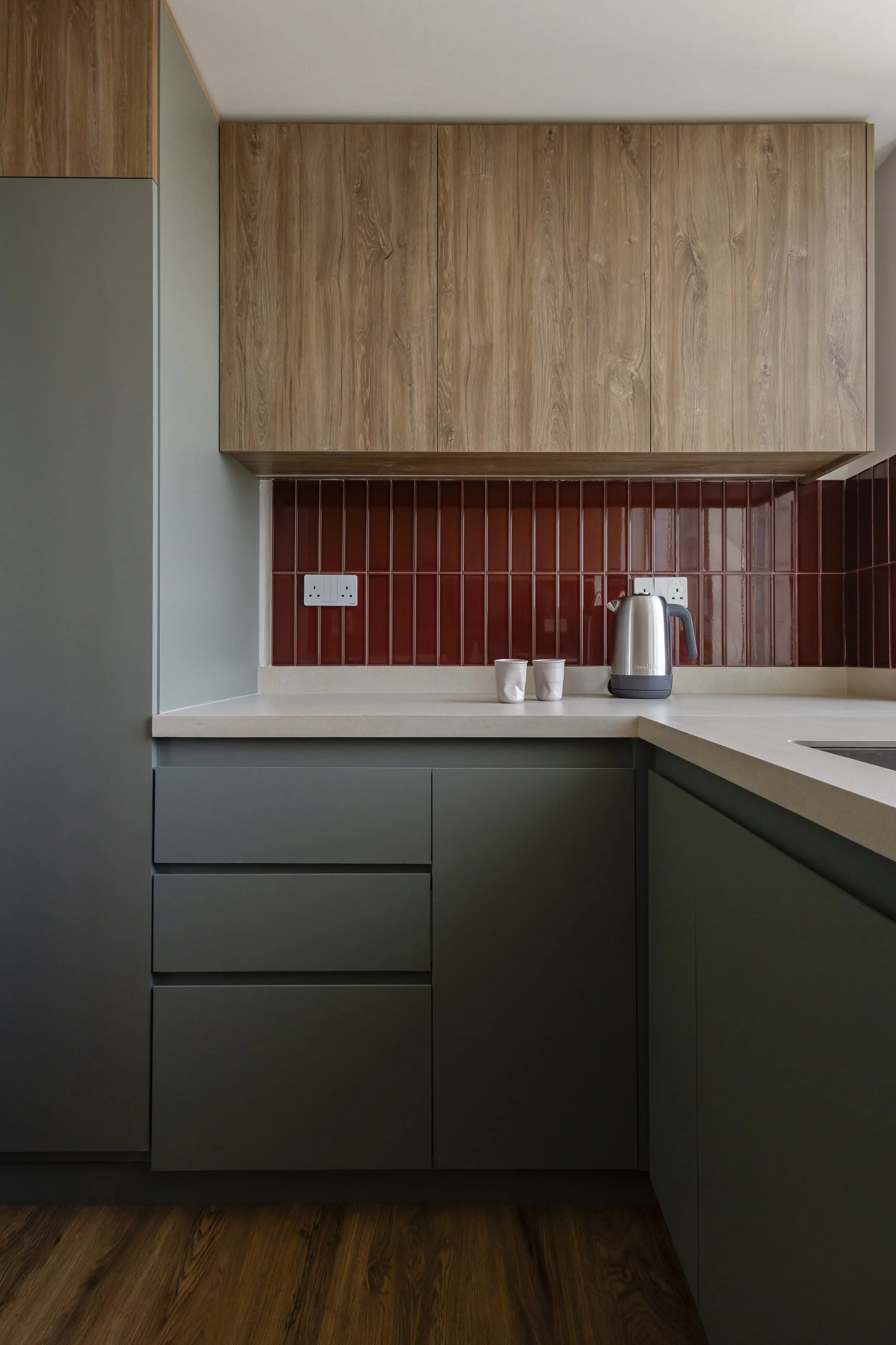 Modern kitchen with green lower cabinets, wooden upper cabinets, red tile backsplash, a beige countertop, a stainless steel electric kettle, and two white cups.