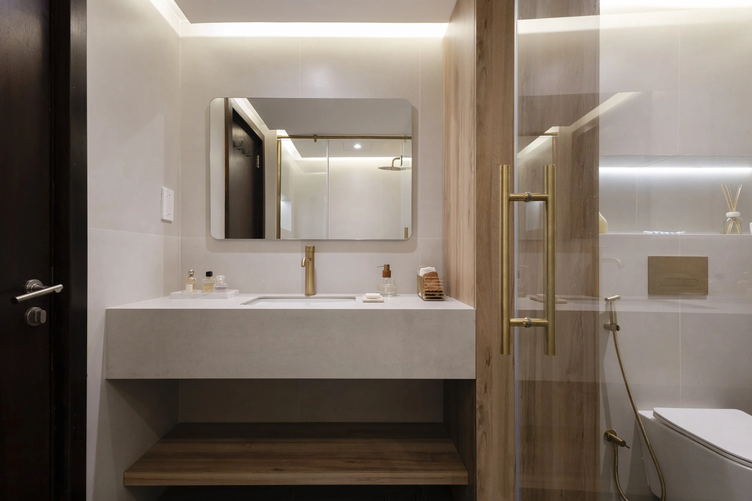 Modern bathroom with a white vanity, a large mirror, gold fixtures, and a glass shower door. There is a toilet with a bidet attachment visible through the glass.