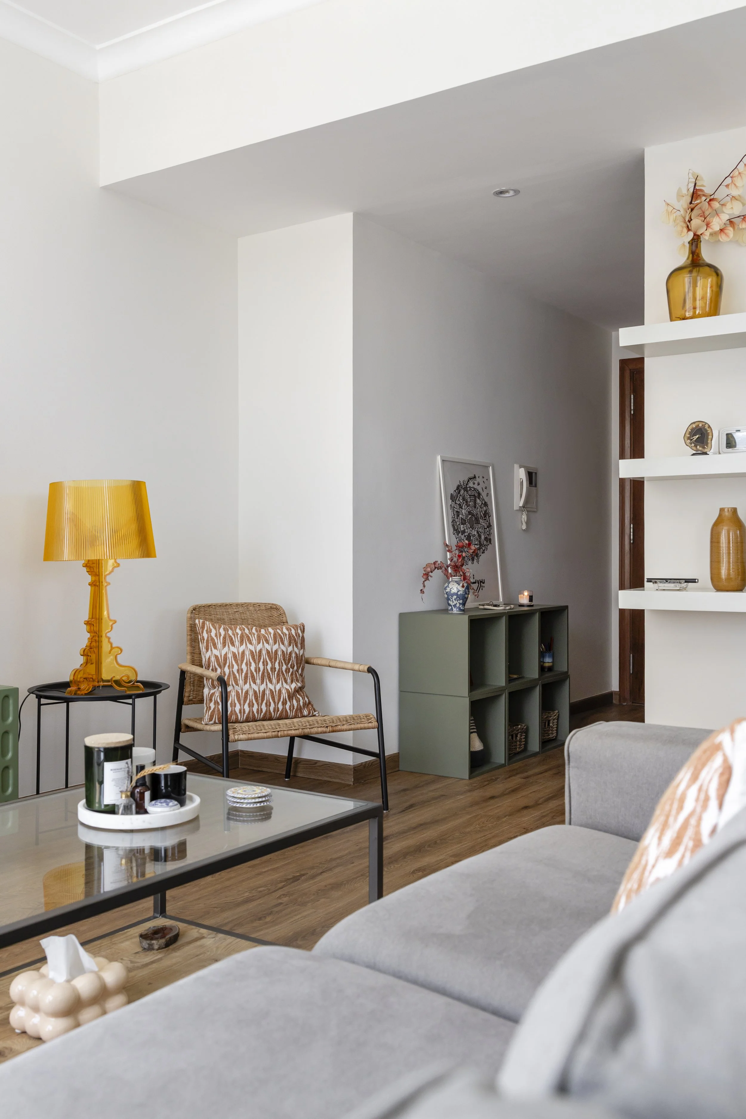 Living room with a gray sofa, coffee table, beige wicker chair with a patterned cushion, yellow lamp, and decorative shelves with vases, artwork, and small decor items.