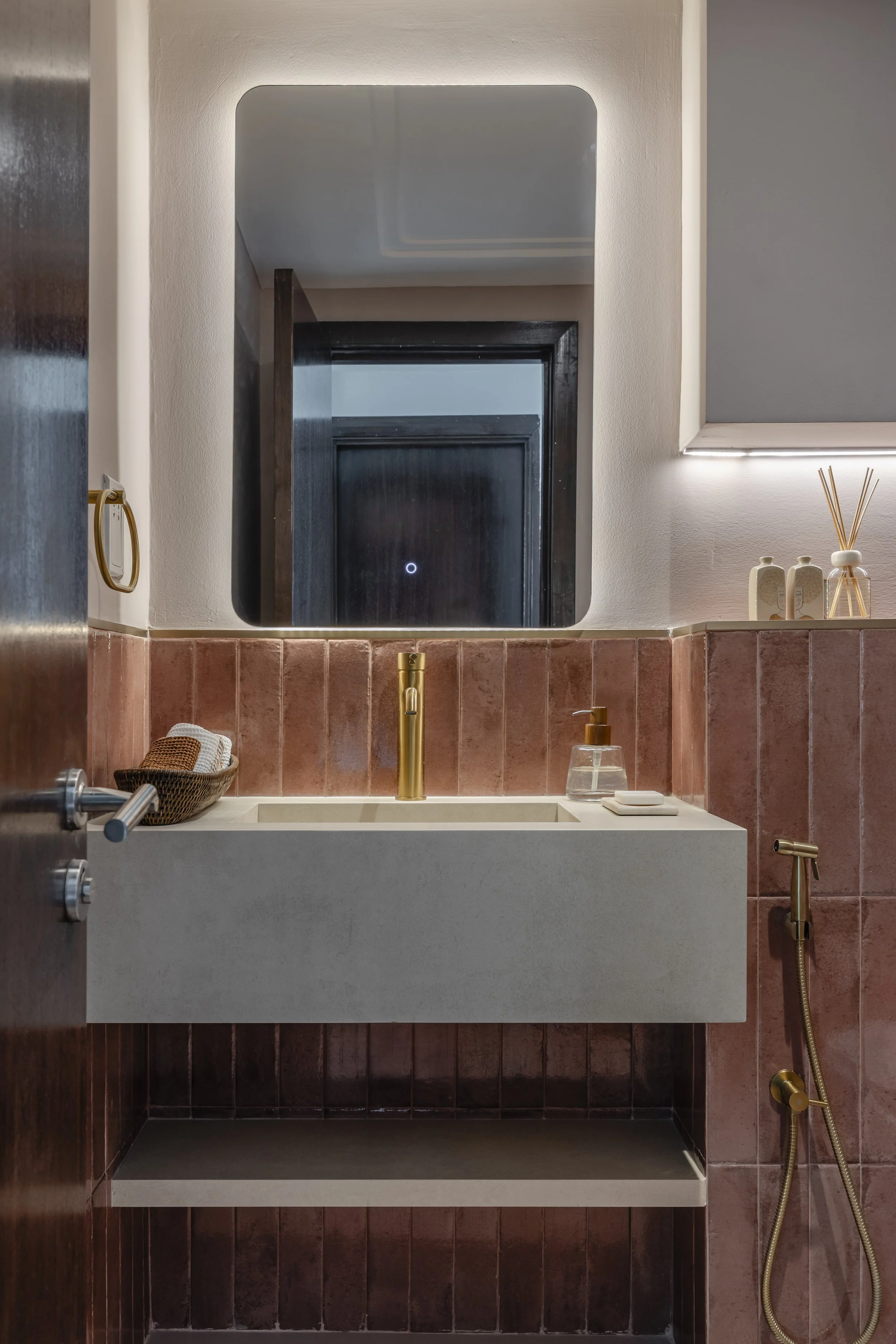 Modern bathroom with a rectangular sink, gold faucet, pink tiled wall, and a mirror reflecting dark door frame.