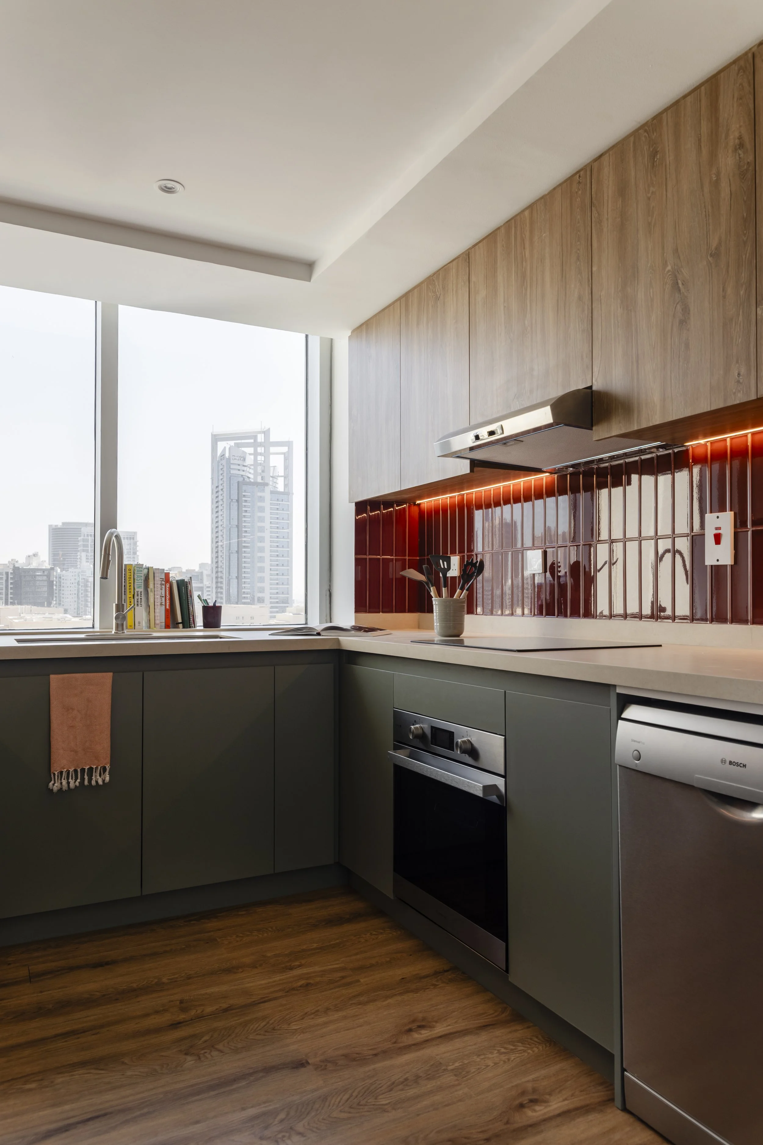 Modern kitchen with dark green lower cabinets, wooden upper cabinets, and a red tiled backsplash, overlooking a city skyline through large windows.