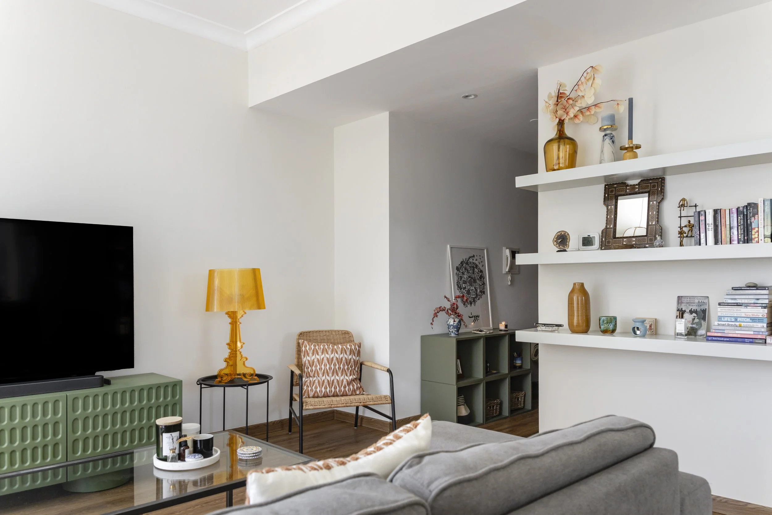 Living room with beige walls, a gray sofa, a green entertainment center, a yellow table lamp, a rattan chair, and white floating shelves with decorative items and books.