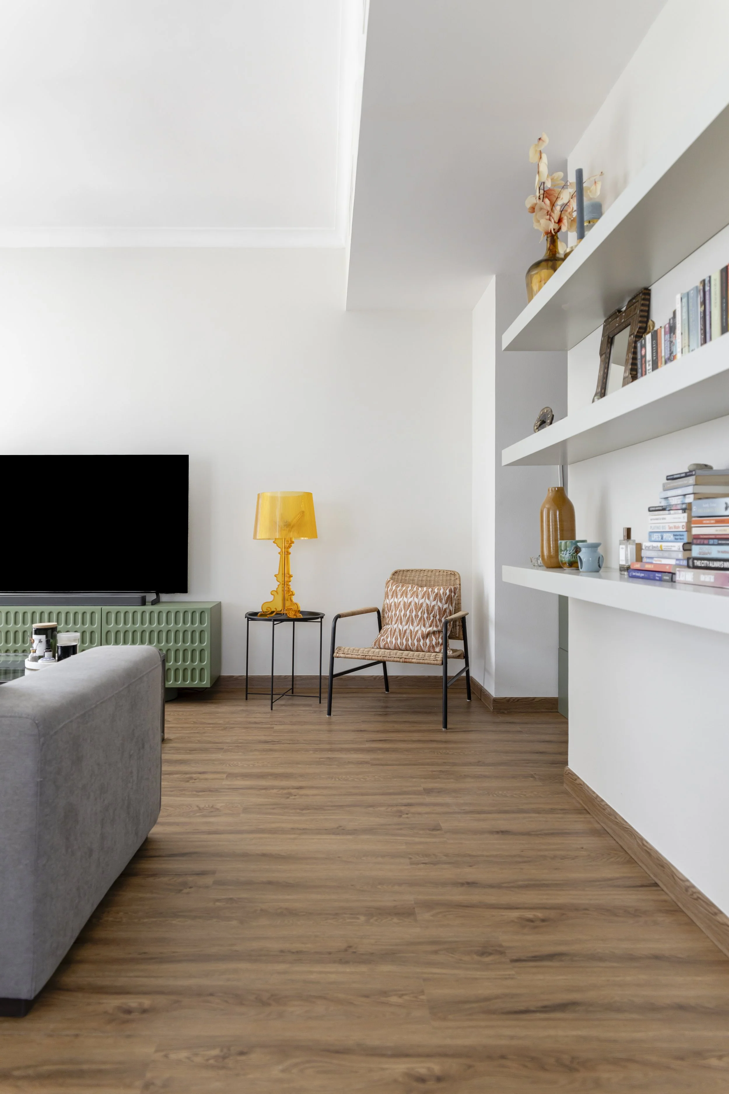 Living room with hardwood floor, white walls, a TV on a green cabinet, a yellow lamp, a patterned armchair, and white shelves with books and decorative vases.