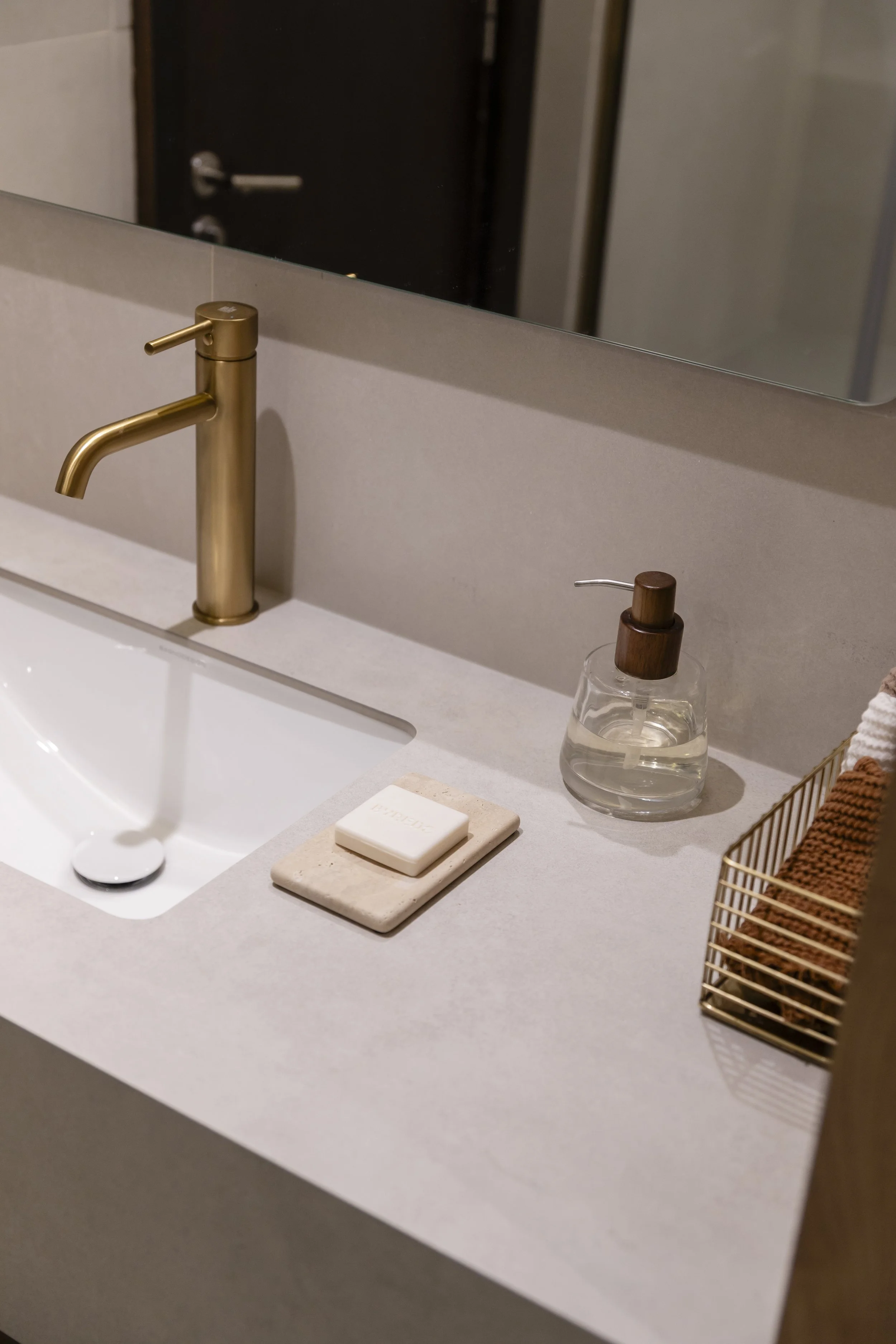 Bathroom sink with gold faucet, soap dispenser, soap on a beige tile, and a gold wire basket holding brown towels, reflected in a mirror.