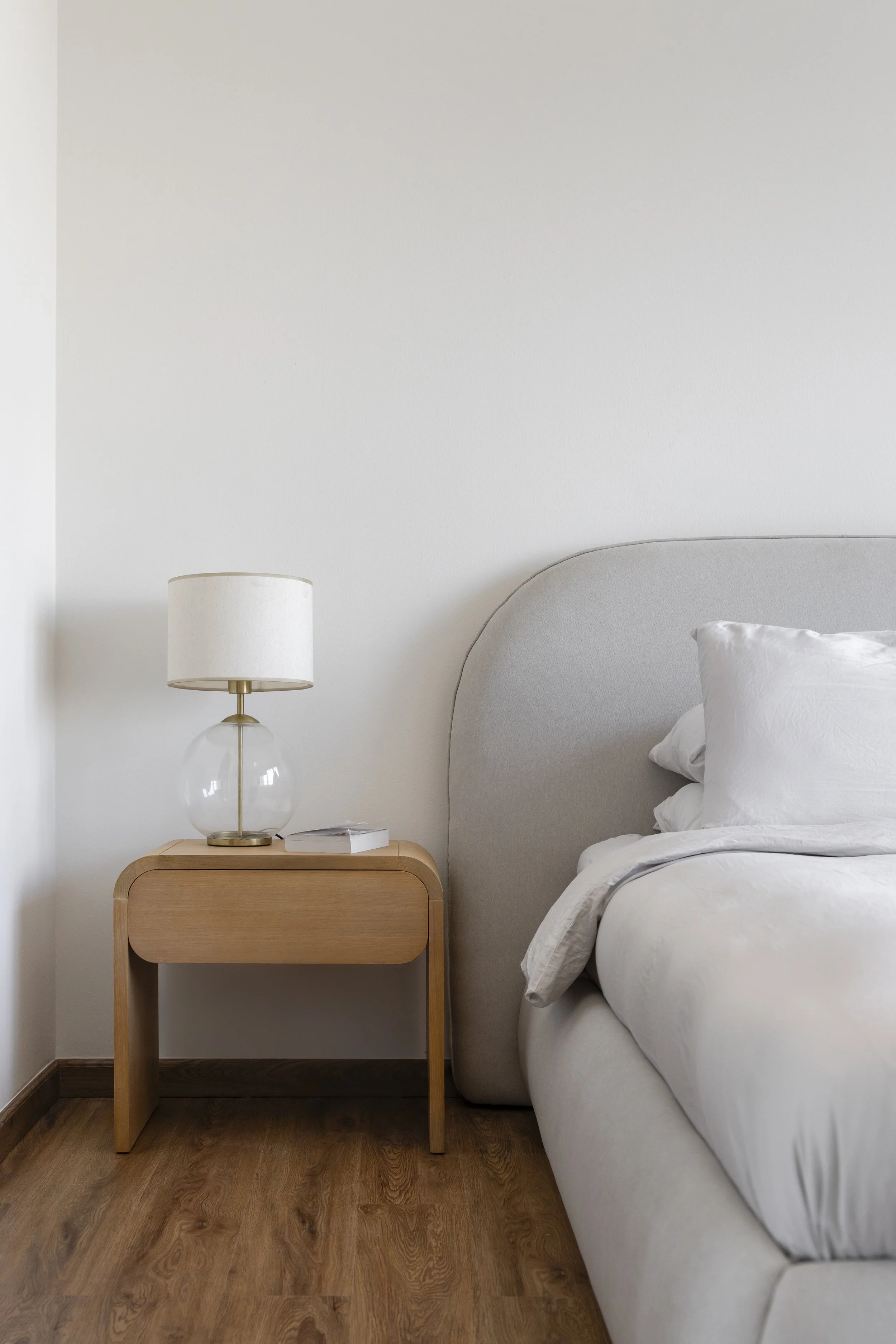 A bedroom with a nightstand, a lamp, a book, and a bed with white bedding and pillows. The room has a wood floor and a plain white wall.