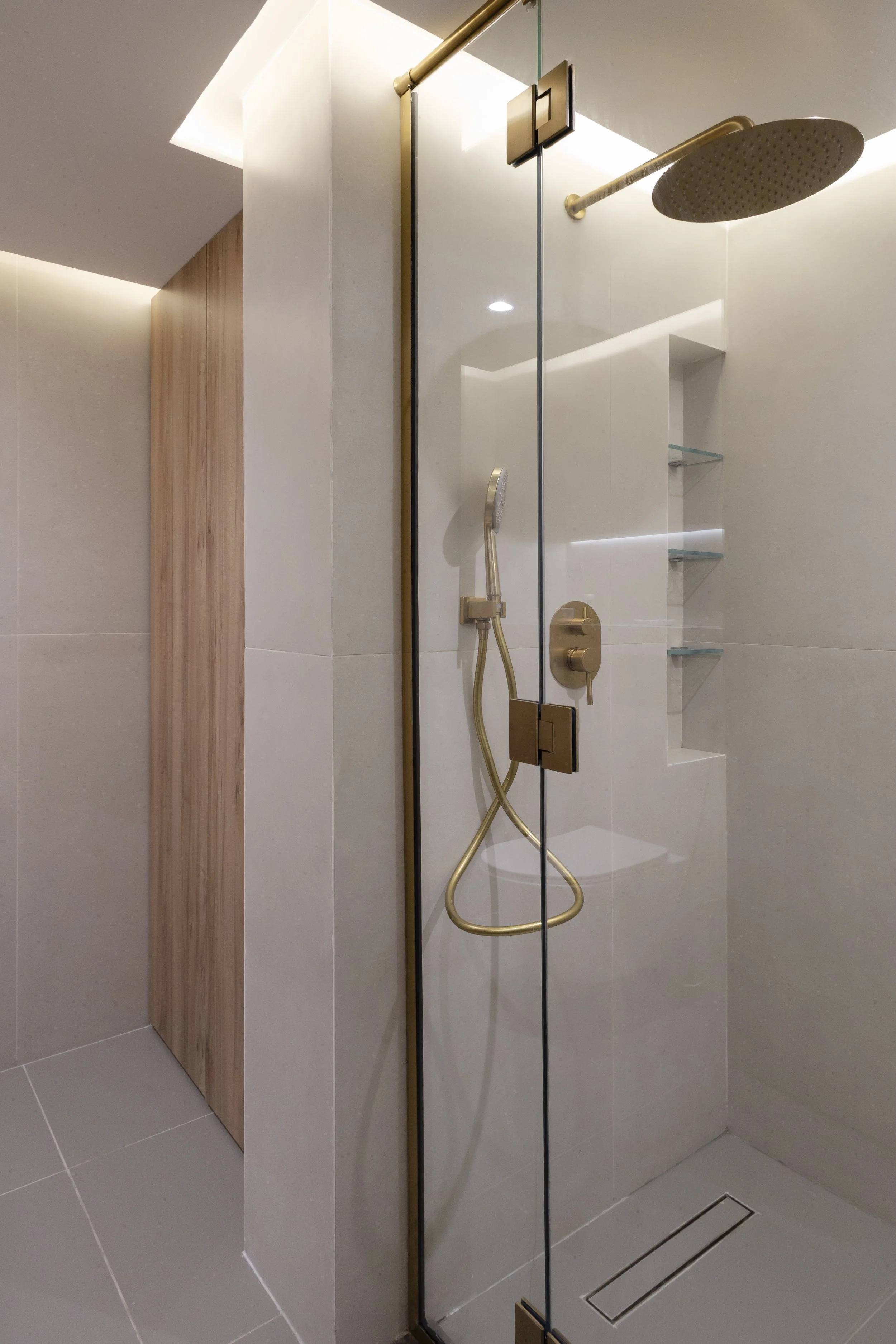 Modern shower with gold fixtures, handheld showerhead, overhead rain shower, glass door, built-in shelves, gray tiled floor, and beige tiled walls.