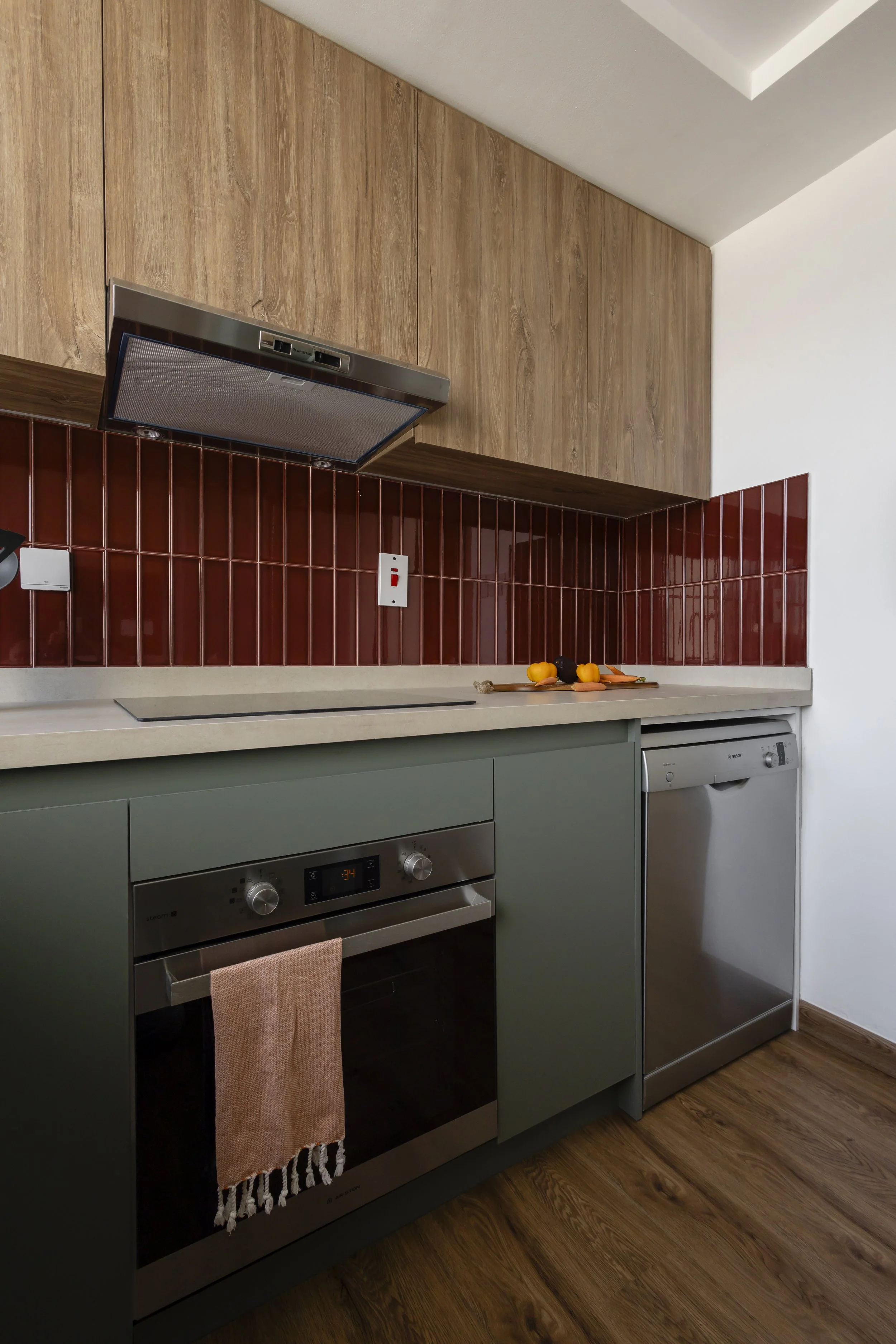 Modern kitchen with green lower cabinets, red backsplash tiles, wooden upper cabinets, a built-in oven, and a dishwasher. There are some vegetables and a cutting board on the countertop.