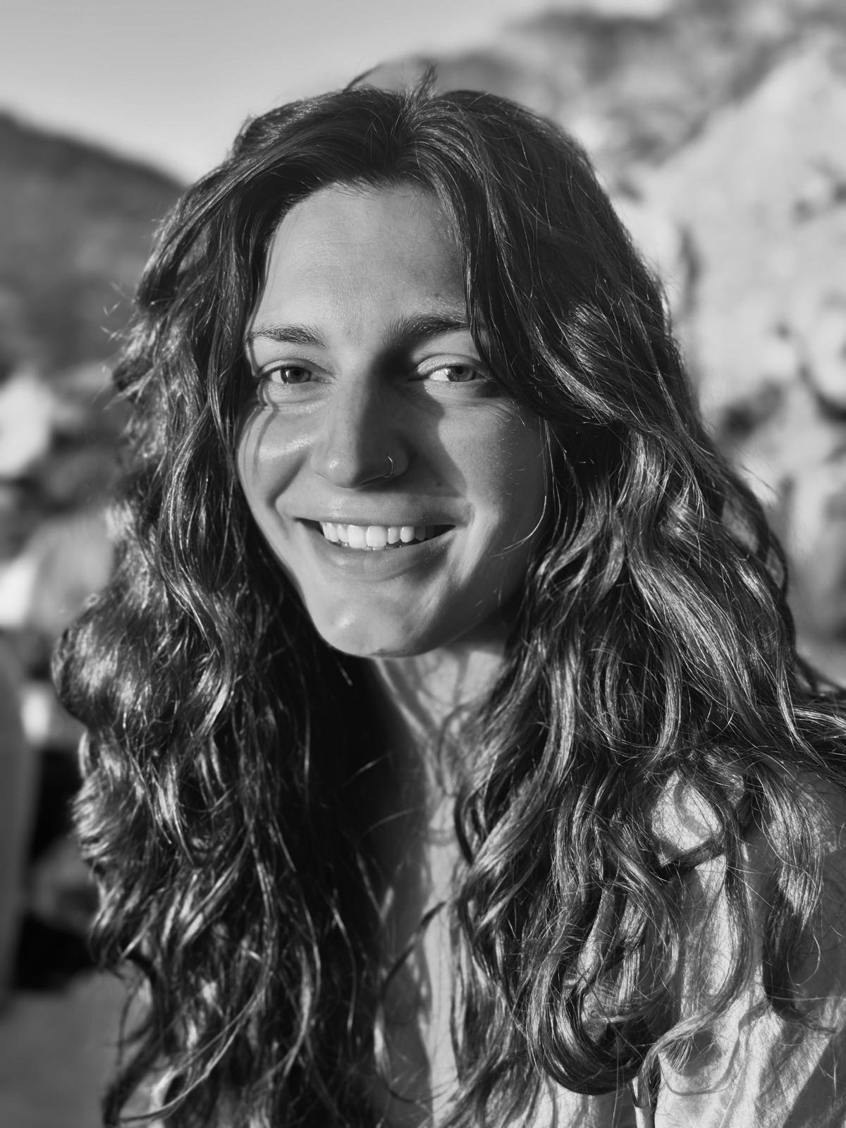 Black and white photo of a smiling woman with long, curly hair and a nose ring, outdoor background.