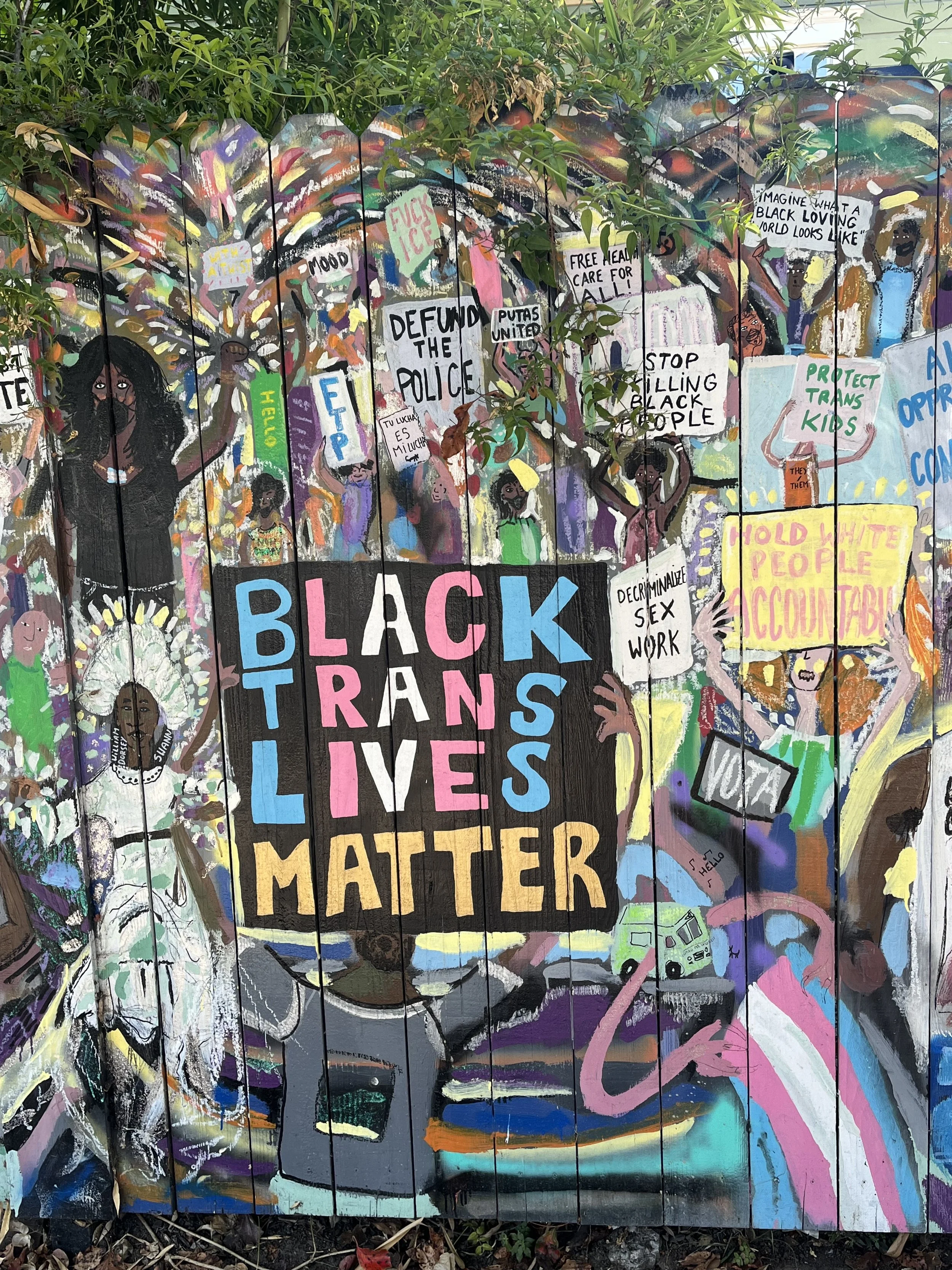 Image description: a photograph of graffiti of a protest with black figures carrying signs saying, “Black trans lives matter”, “protect trans kids” and “decriminalise sex work”.
