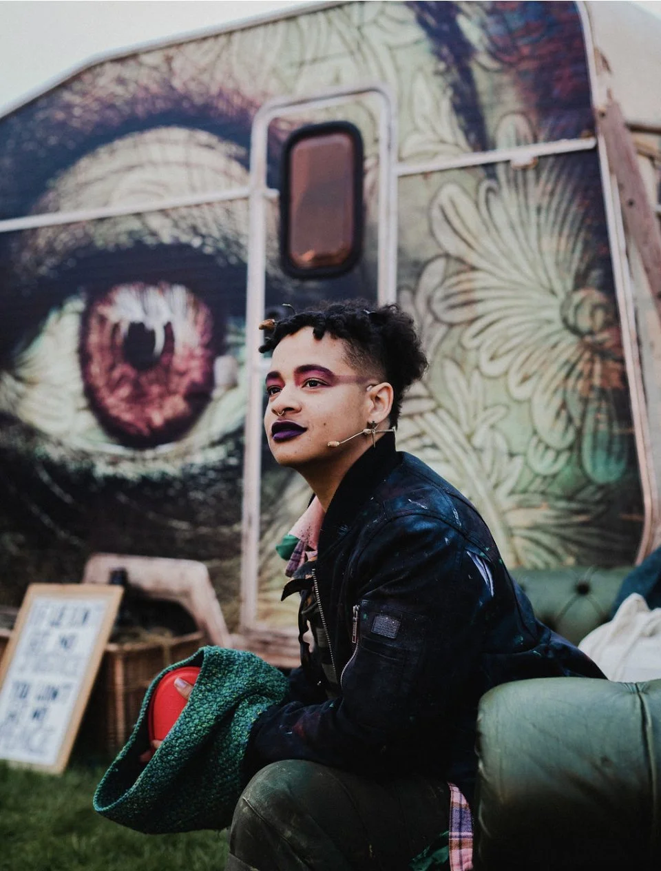 Image description: Elk in character as a nymph from Galatea, a site-specific theatre  produced as part of Brighton Festival. They wear tattered clothes, dramatic makeup and sit in front of an intricately graffiti’d caravan.
