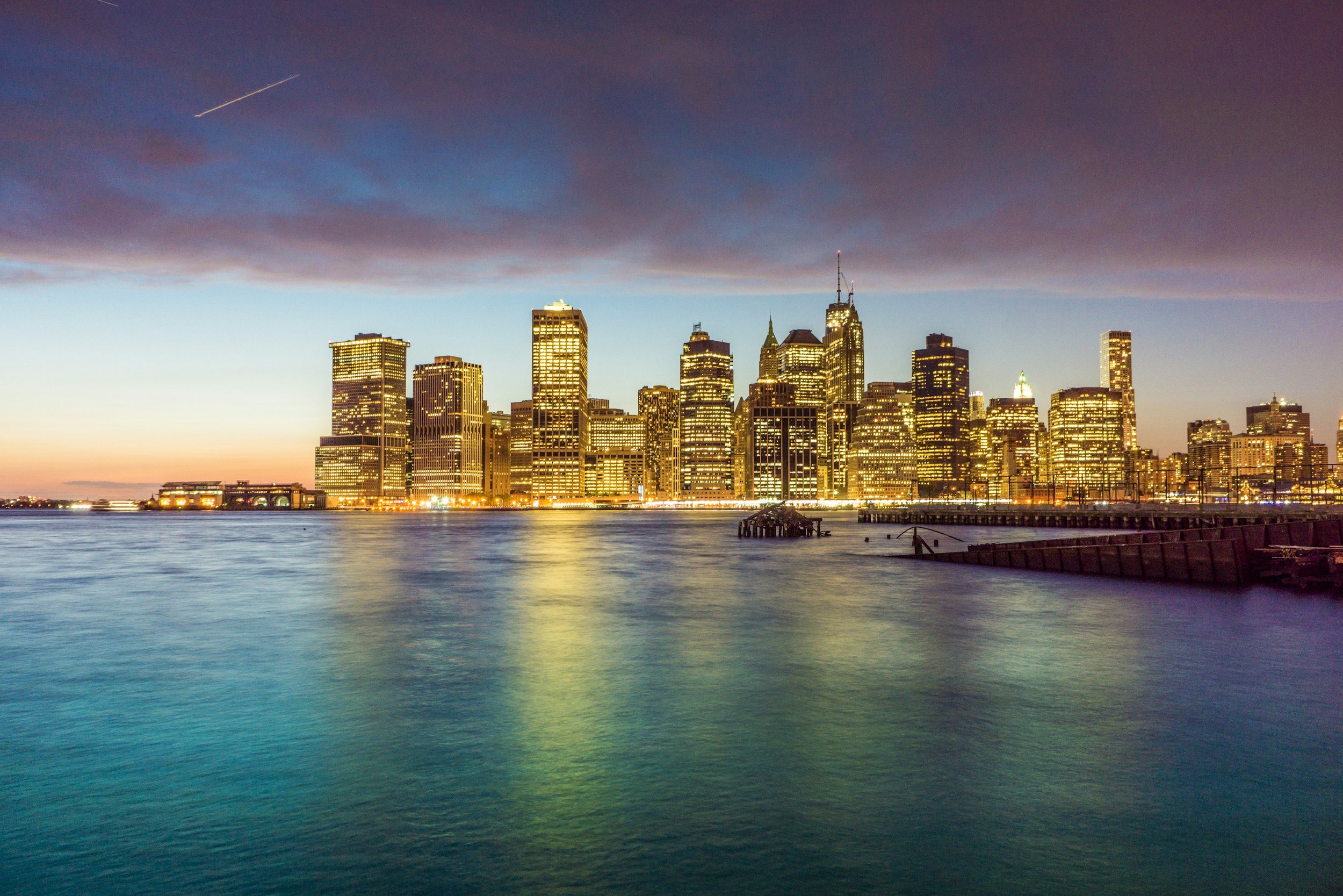 New York City skyline at dusk with illuminated skyscrapers and water in the foreground.