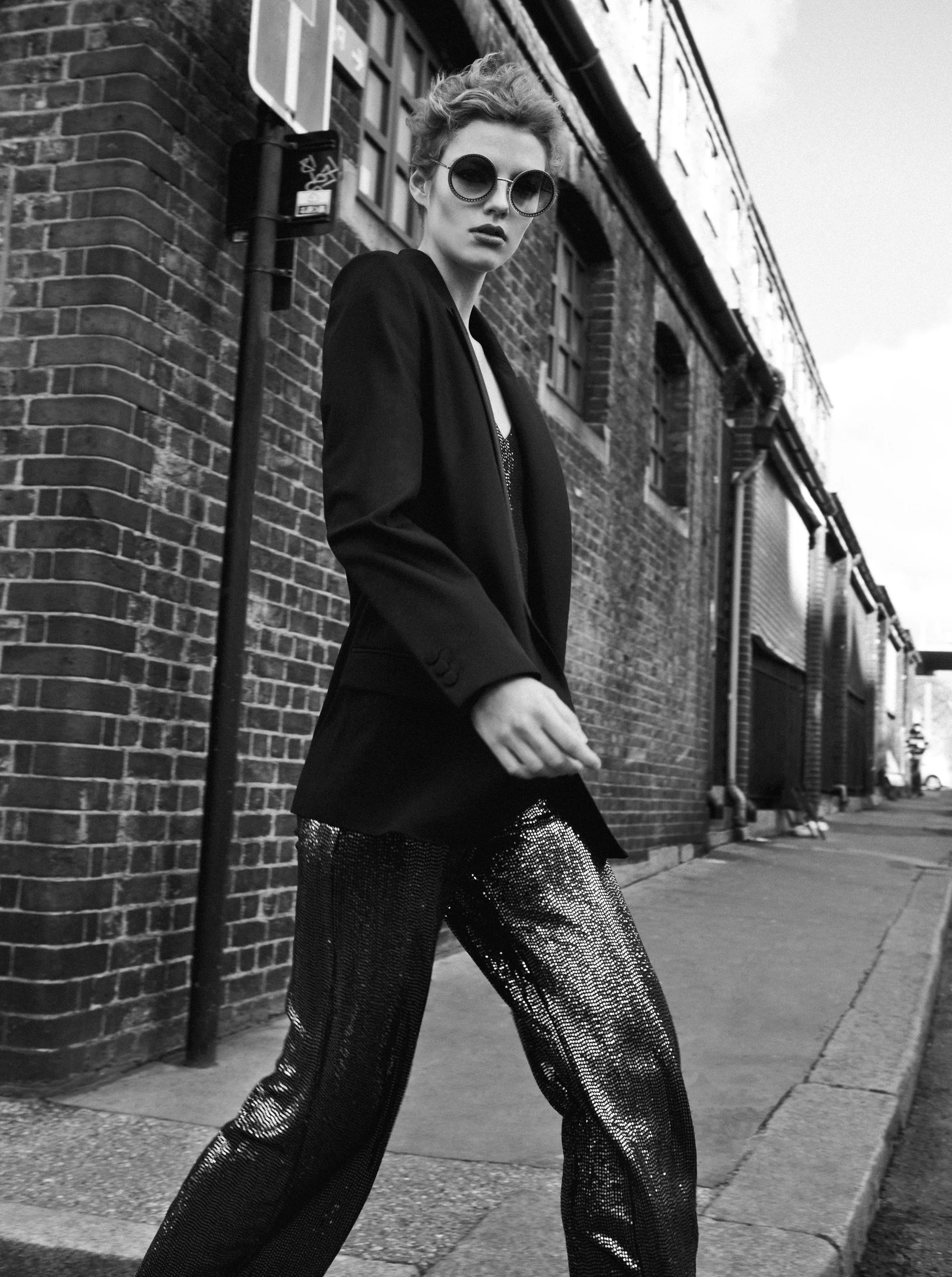 A woman wearing large round sunglasses, a dark blazer, and shimmering wide-leg pants, stands on a city sidewalk against a brick building with windows and a streetlight, captured in black and white.