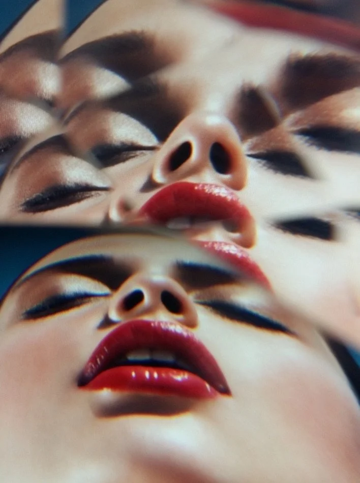 Close-up of a mirrored image of a woman's face with bold makeup and red lipstick, showing multiple overlapping reflections.