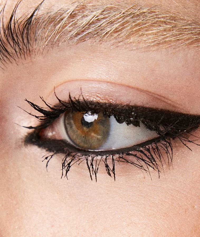 Close-up of a person's eye with makeup including black eyeliner and mascara, brown iris, and blonde eyebrow.