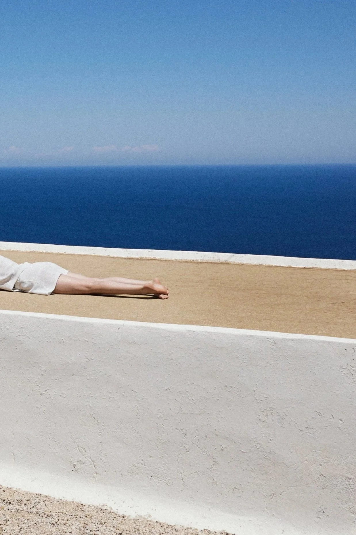 A person lying down on a beige surface with a background of the blue sea and sky, viewed from the side showing their extended arm and part of their leg.