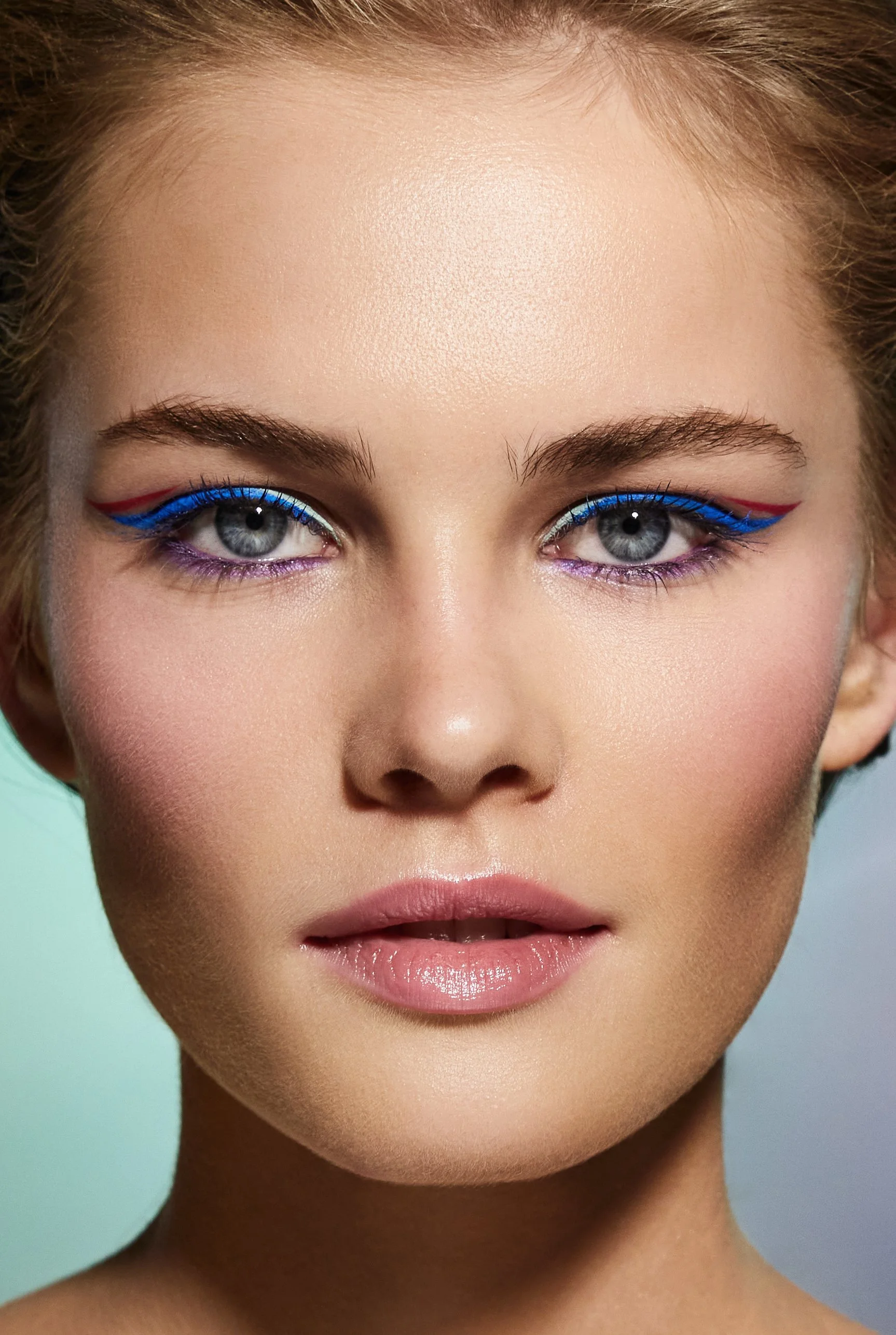 Close-up of a woman's face with bold blue and red eyeliner on her upper eyelids and purple eyeliner on her lower eyelids, with natural makeup and neutral lips.