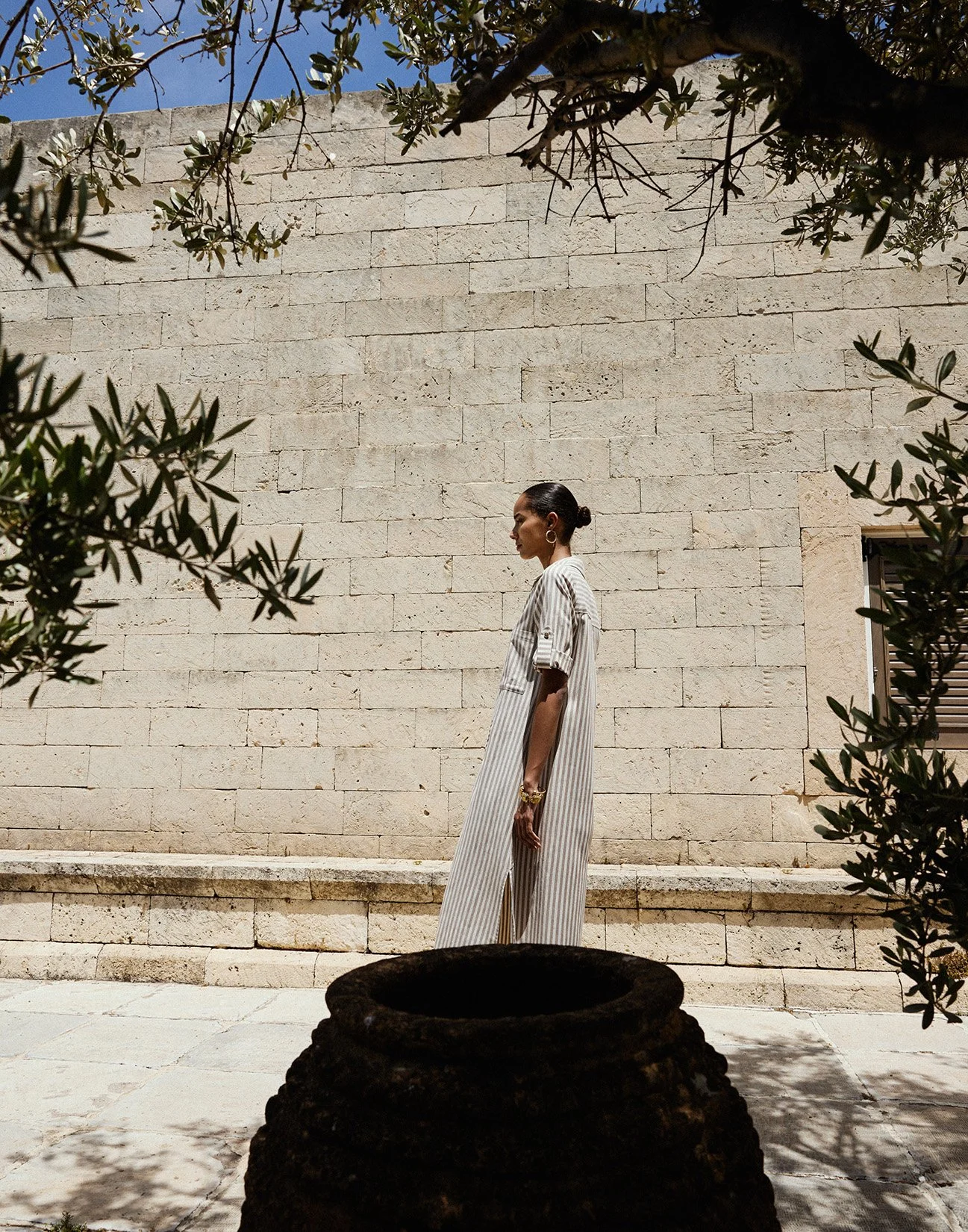 A woman in a striped dress stands against a beige brick wall, framed by leafy branches in the foreground, under a clear blue sky.