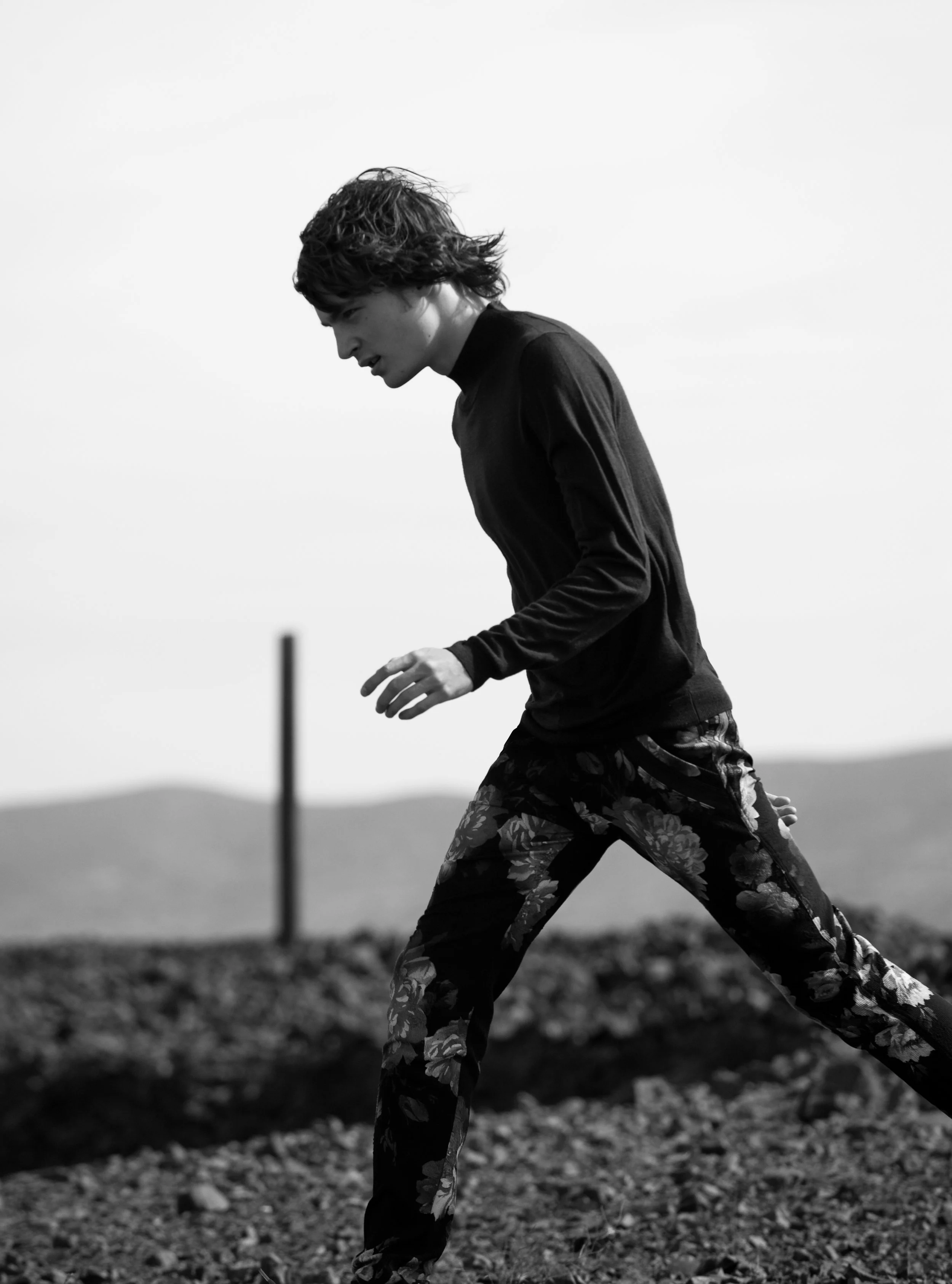 A young person running outdoors on a rocky terrain, wearing dark clothing and patterned pants, captured in black and white with a blurred background.