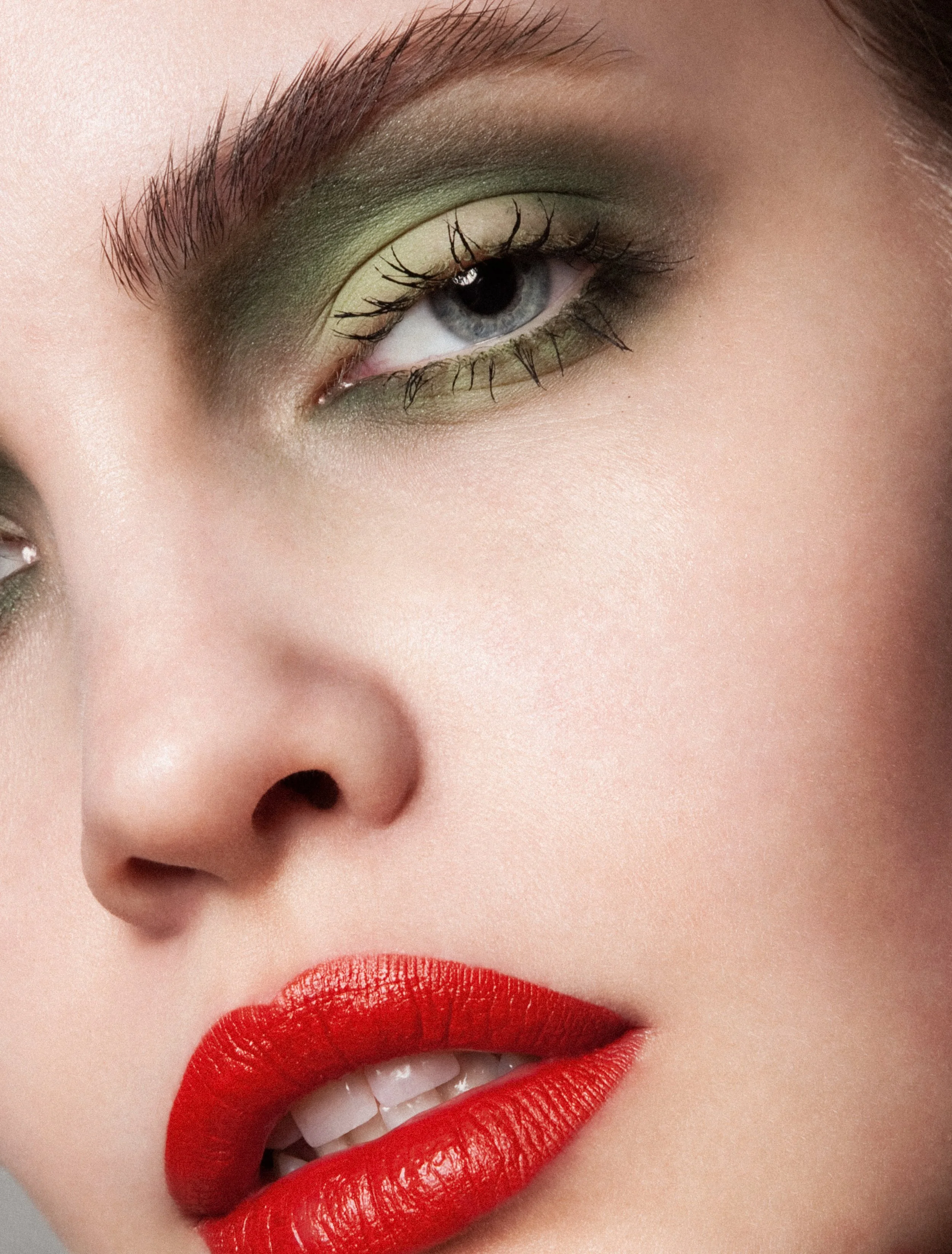 Close-up of a woman's face showing her eye with green eyeshadow, long eyelashes, well-defined eyebrows, red lipstick, and partially visible nose and lips.