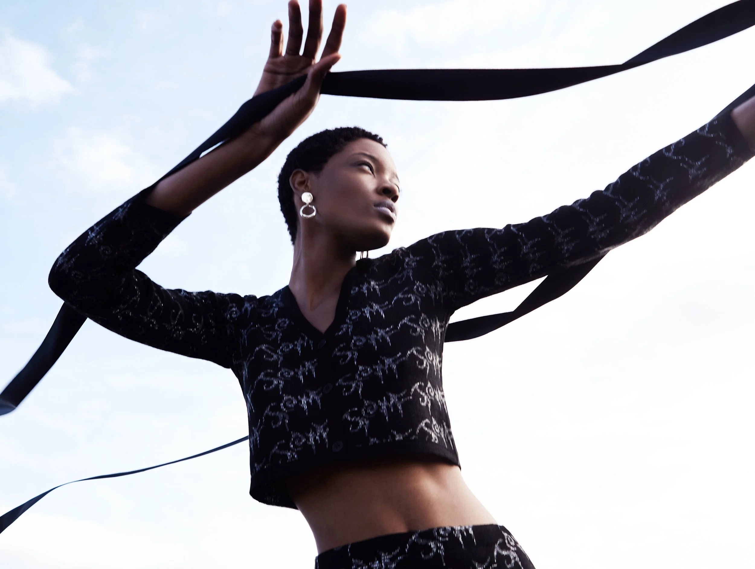 A woman with short hair and earrings is outdoors, lifting black fabric strips above her head against a blue sky with clouds.