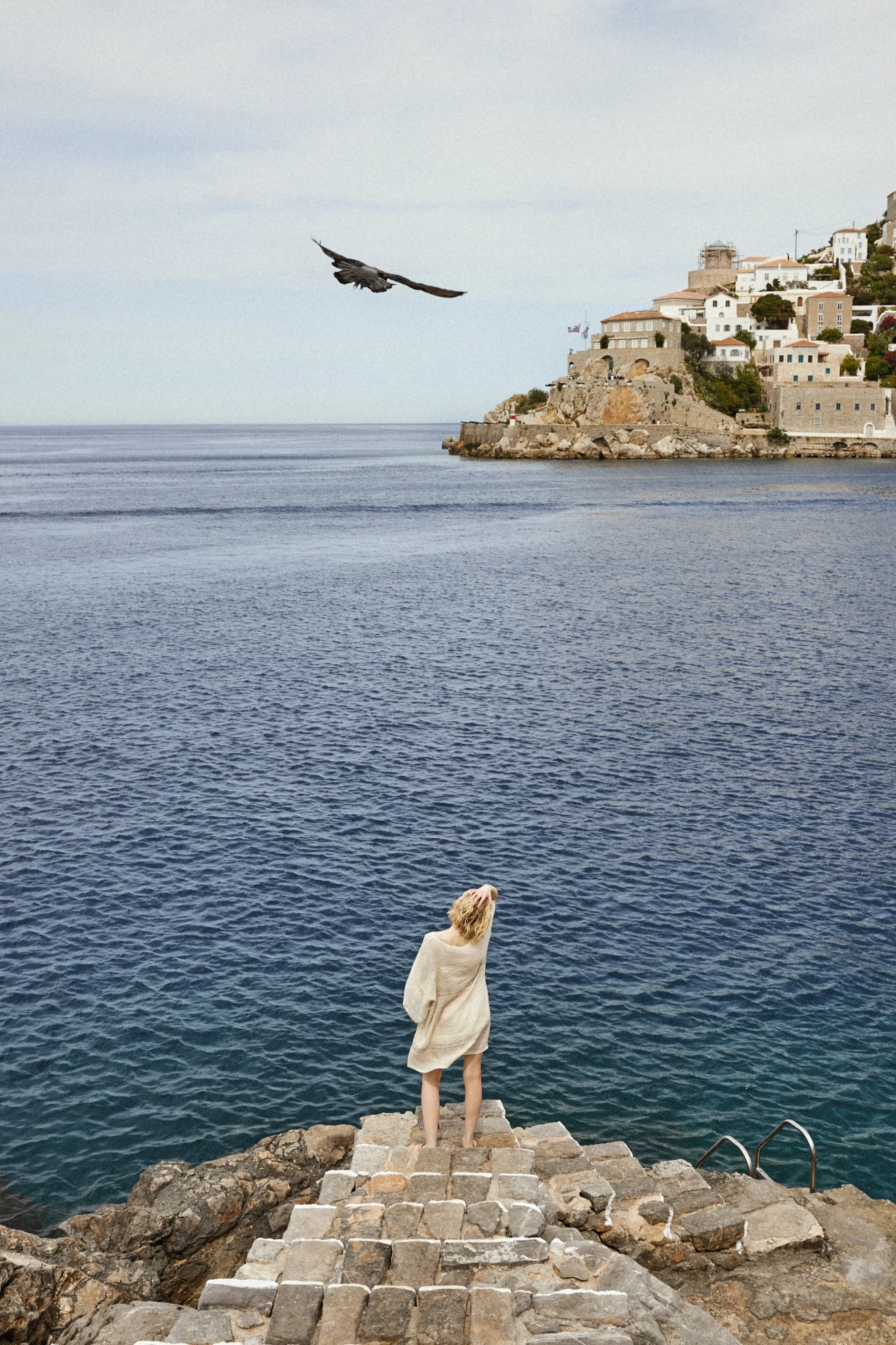 A person with blonde hair, wrapped in a beige towel, standing on a stone pier overlooking the ocean, with a hill of white houses in the background and a seagull flying overhead.