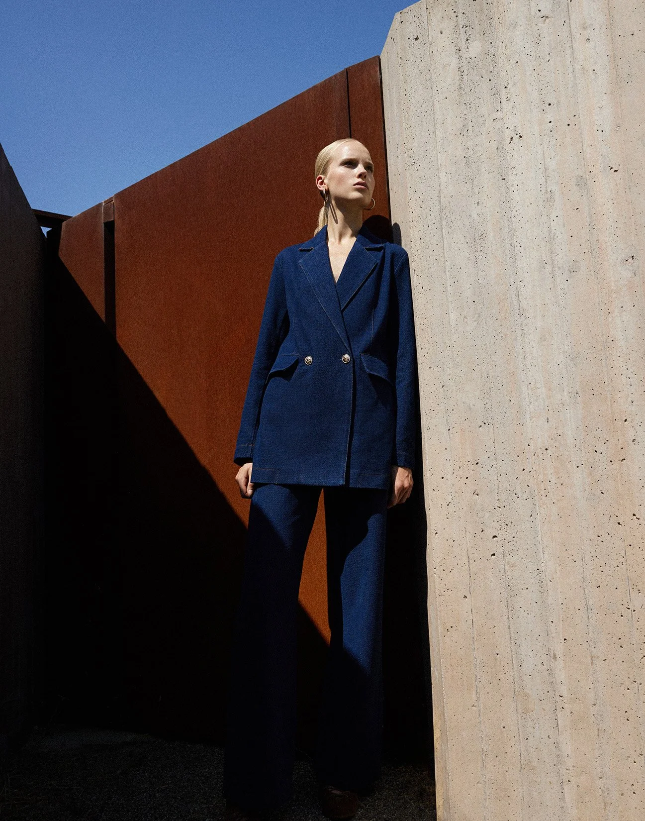 A woman standing outdoors next to a concrete wall and a rust-colored metal panel, dressed in a dark blue suit with a blazer and matching pants, against a bright blue sky.
