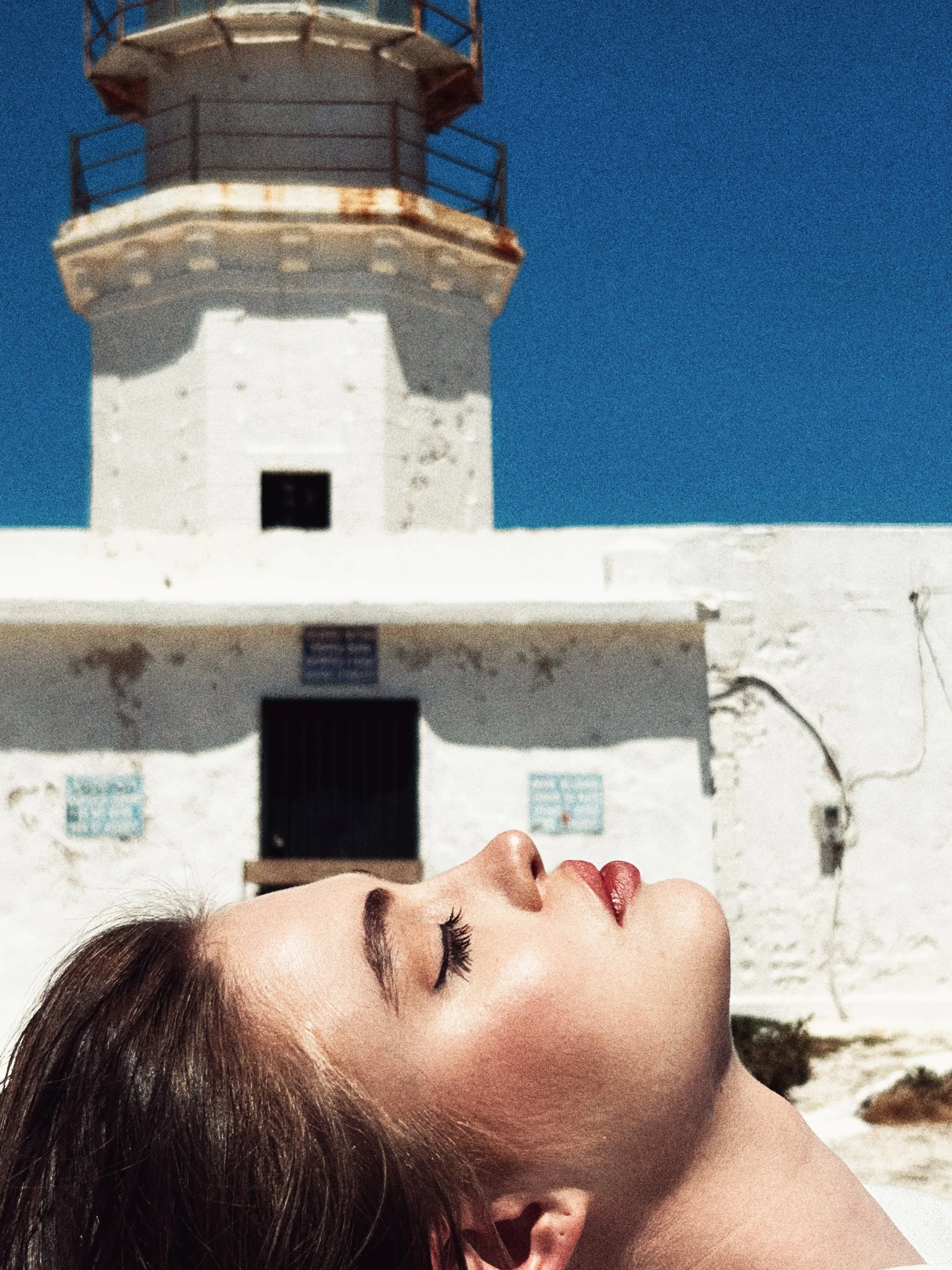 A woman with closed eyes and brown hair lying on her side, with a white building and a lighthouse in the background.