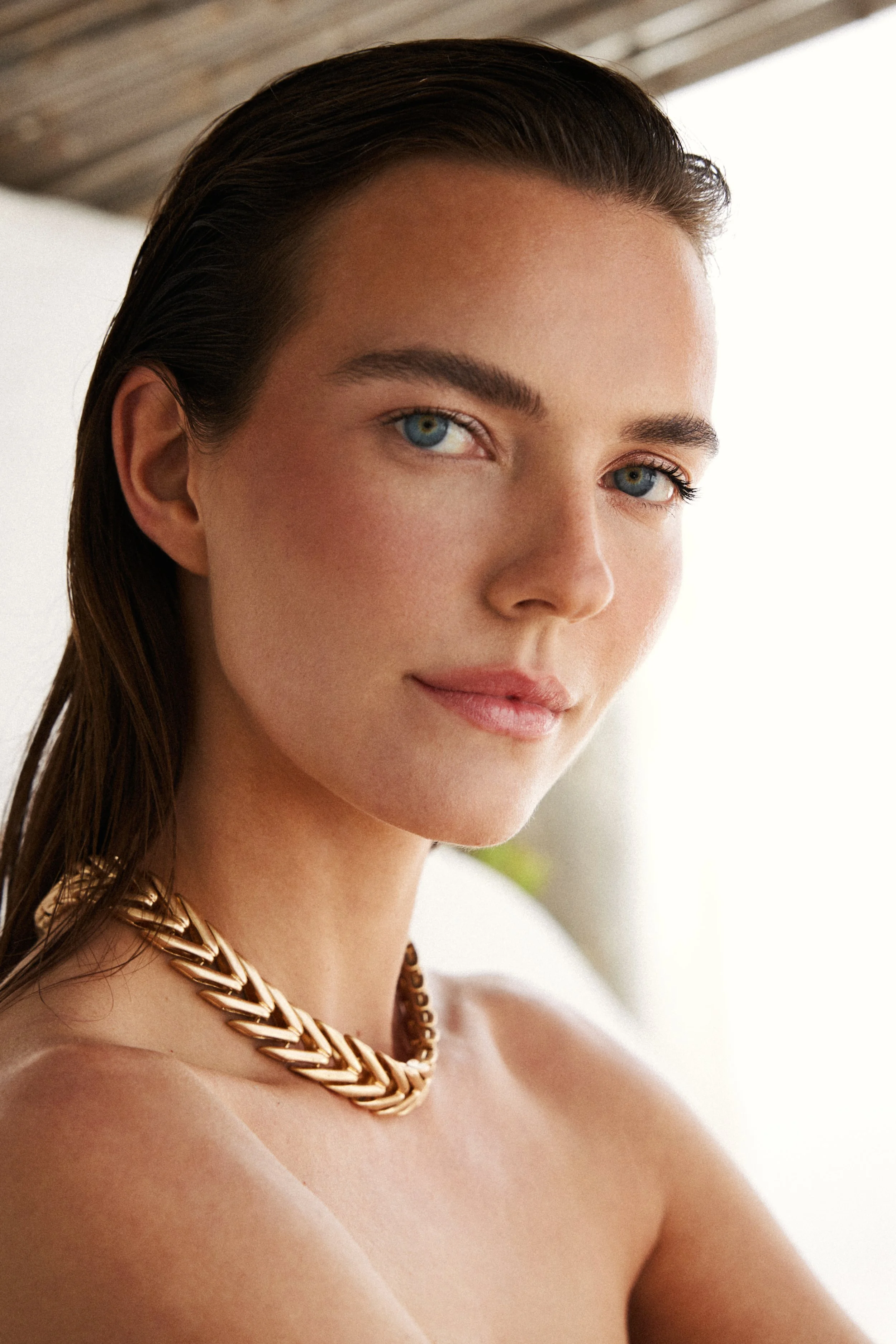 A young woman with blue eyes and dark hair tied back, wearing a gold chain necklace, looking at the camera with a slight smile.