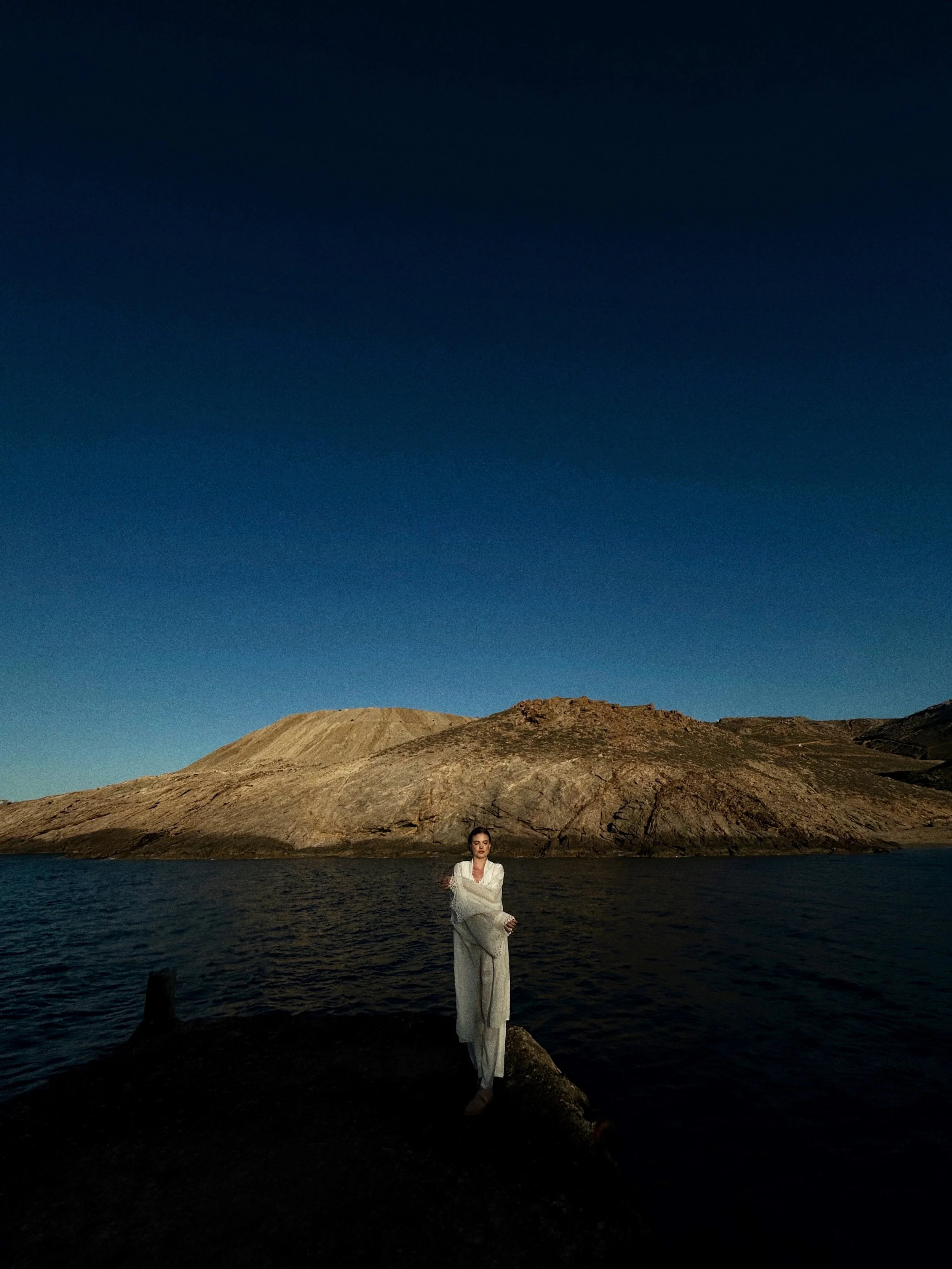 A woman in a white dress standing on a rock by a body of water at dusk, with a mountain in the background and a clear darkening sky above.