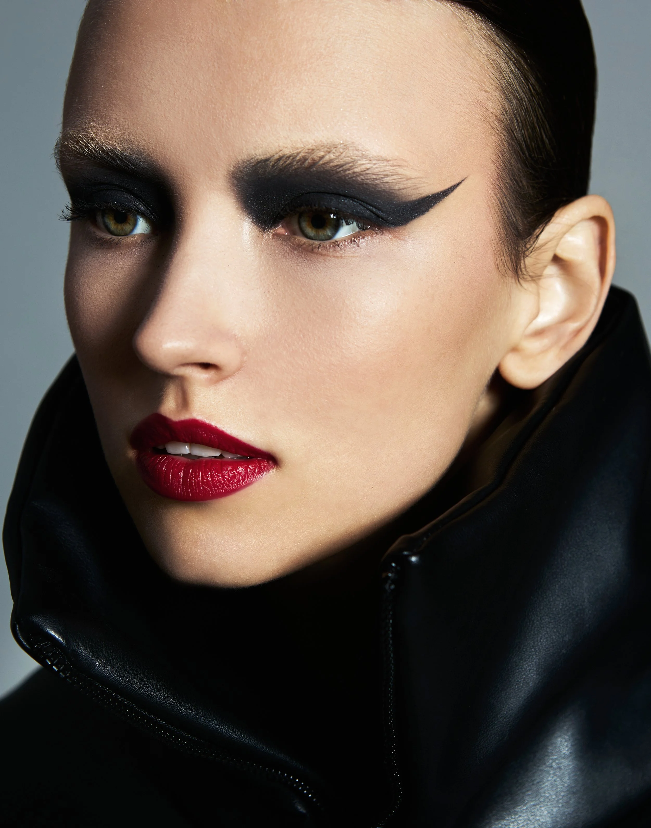 Close-up of a woman with green eyes, bold black eye makeup with a winged eyeliner, red lipstick, and wearing a black leather jacket.