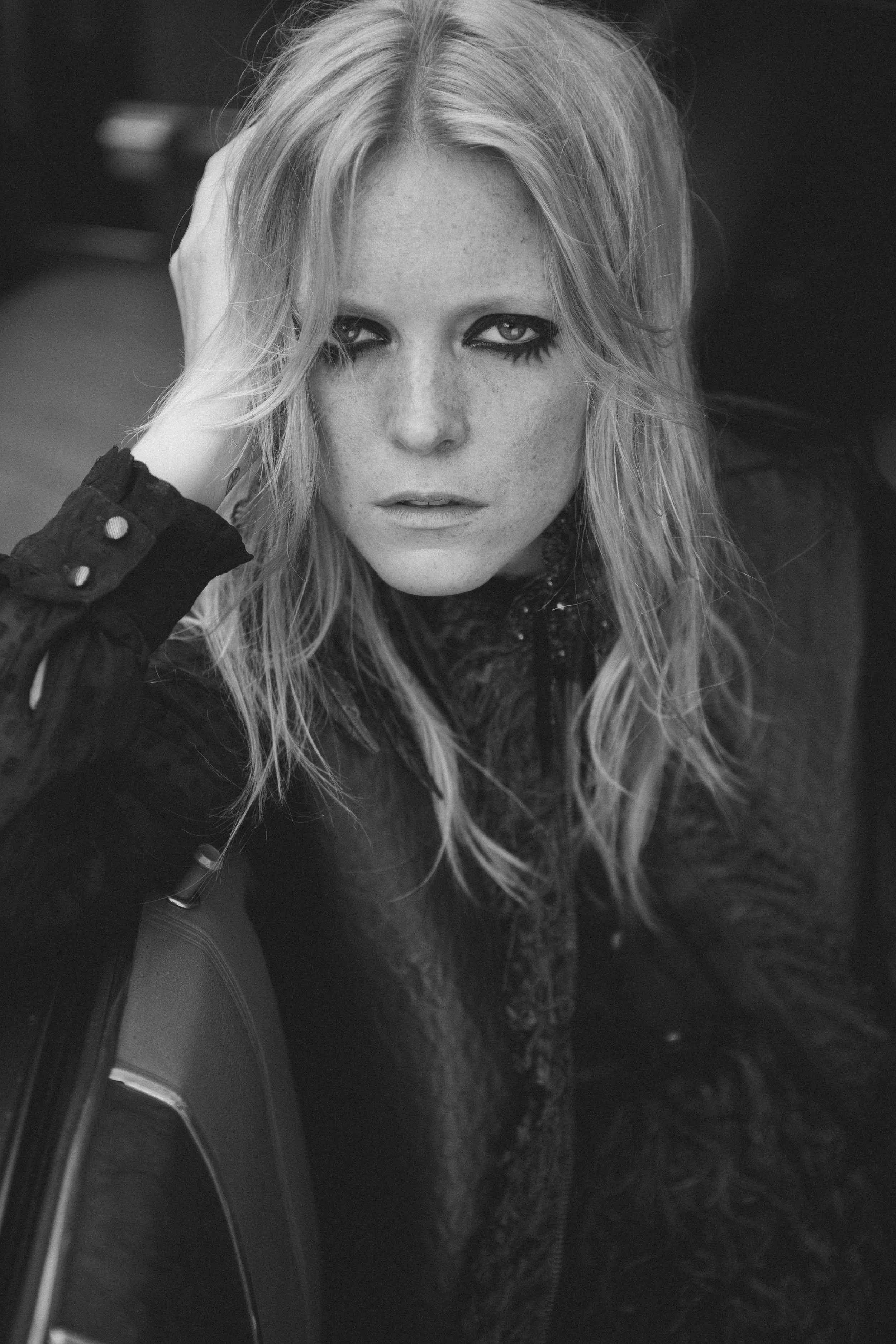 A close-up black and white photo of a woman with long, wavy hair. She has intense eye makeup with dark eyeliner and mascara, with her hand resting on her head, and is wearing a dark, textured jacket.