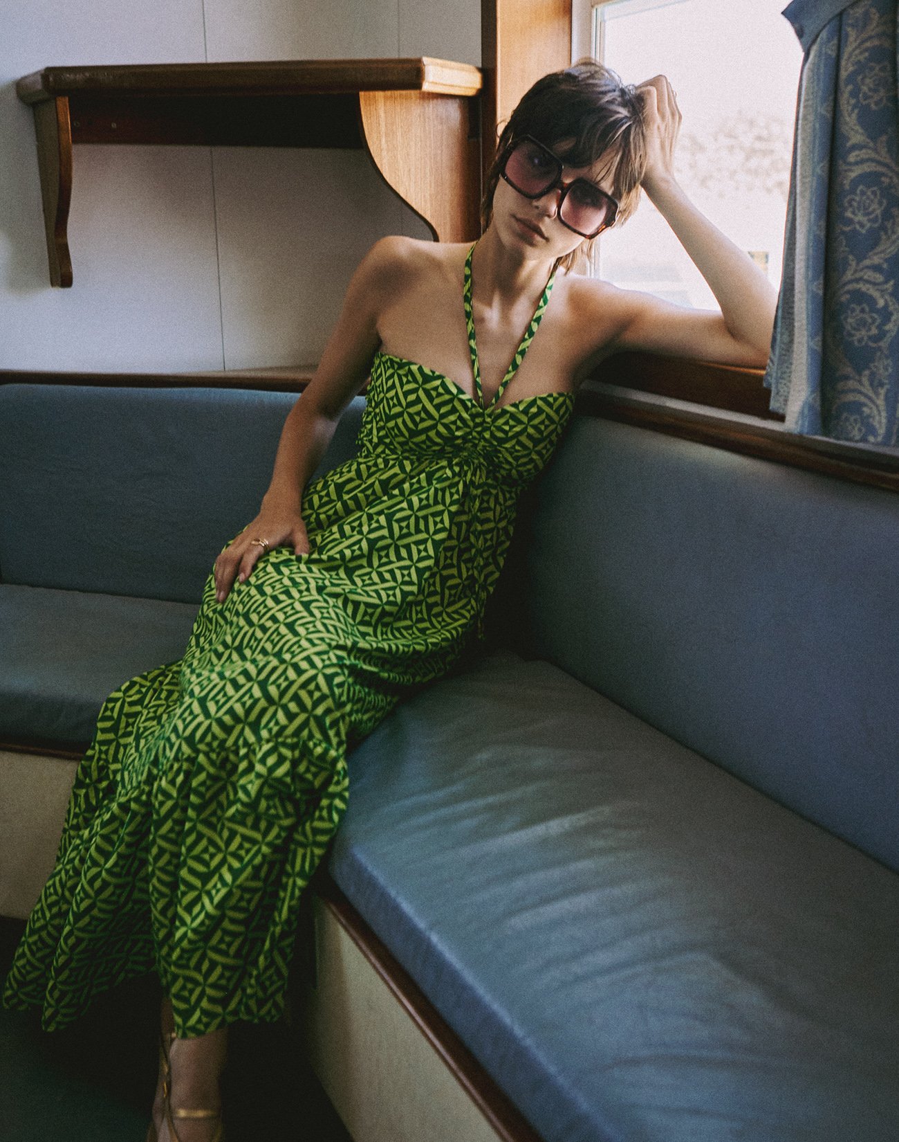Woman sitting on a grey couch by a window, wearing a green geometric patterned dress and sunglasses, with short brown hair, resting her head on her hand.