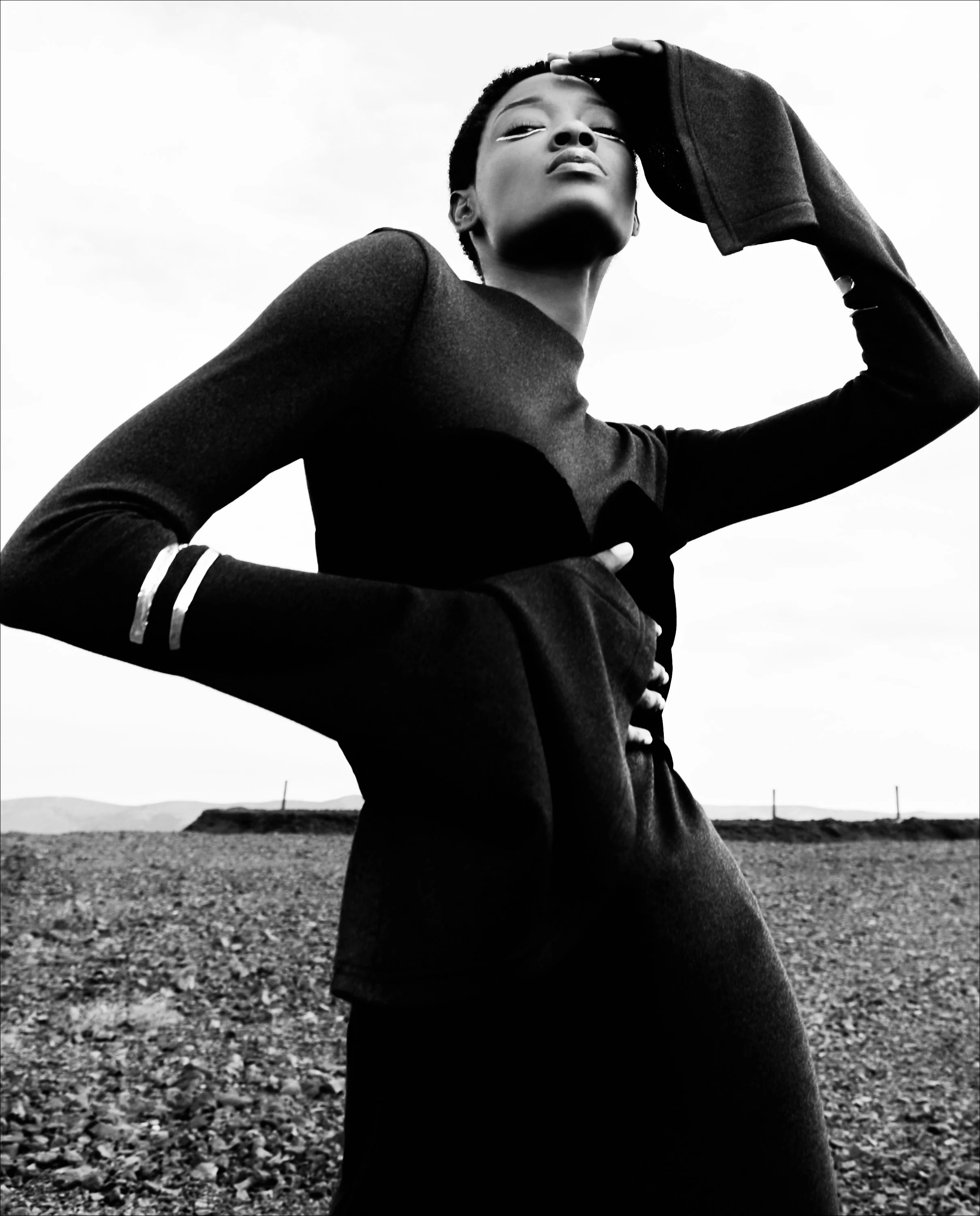A black and white photo of a woman with short hair, wearing a dark long-sleeve top with white stripes on the cuffs. She is outdoors on a rocky terrain with a distant horizon and mountains in the background. She is looking slightly upwards, with one h