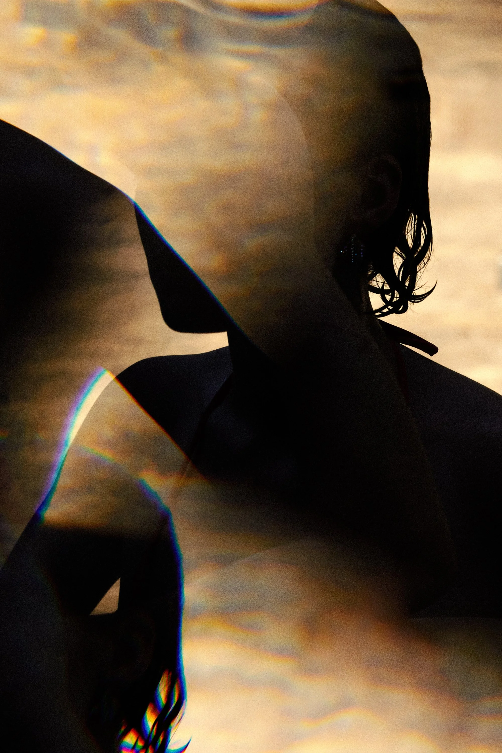 A person with curly hair and earrings, shirtless, is standing with a beach in the background during sunset or sunrise. The scene includes reflections and rainbow-like distortions.