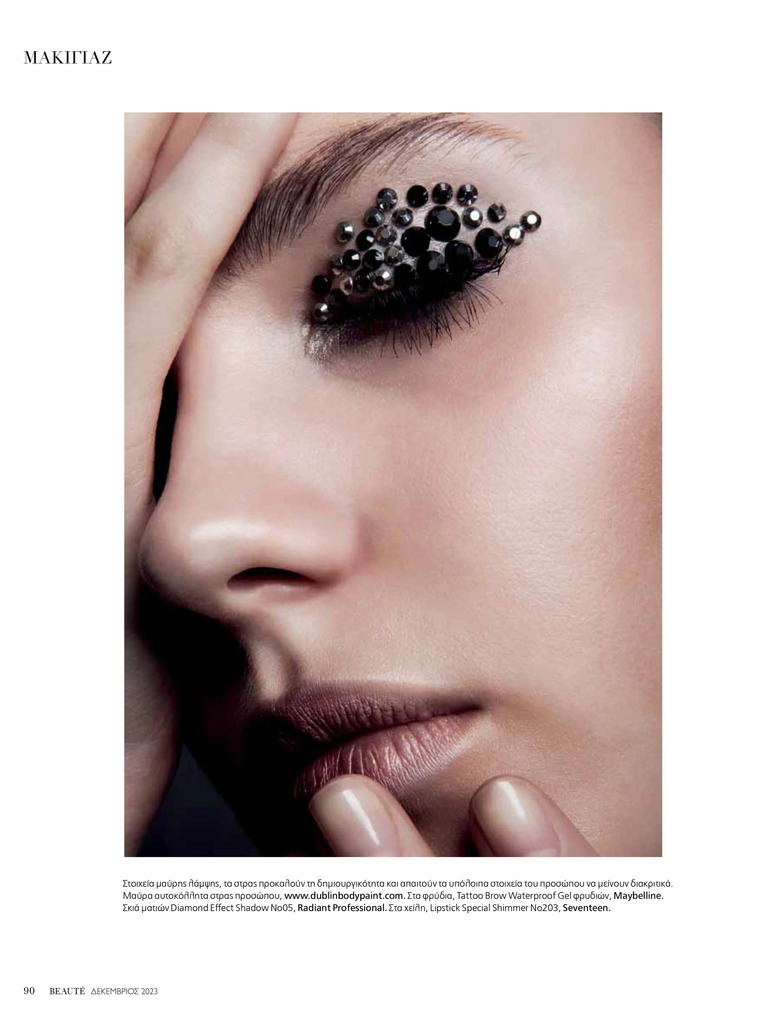 Close-up of a woman's face with a hand touching her face, featuring makeup including black diamond effect shadow, shiny black rhinestones on the eyelid, and brown lipstick.